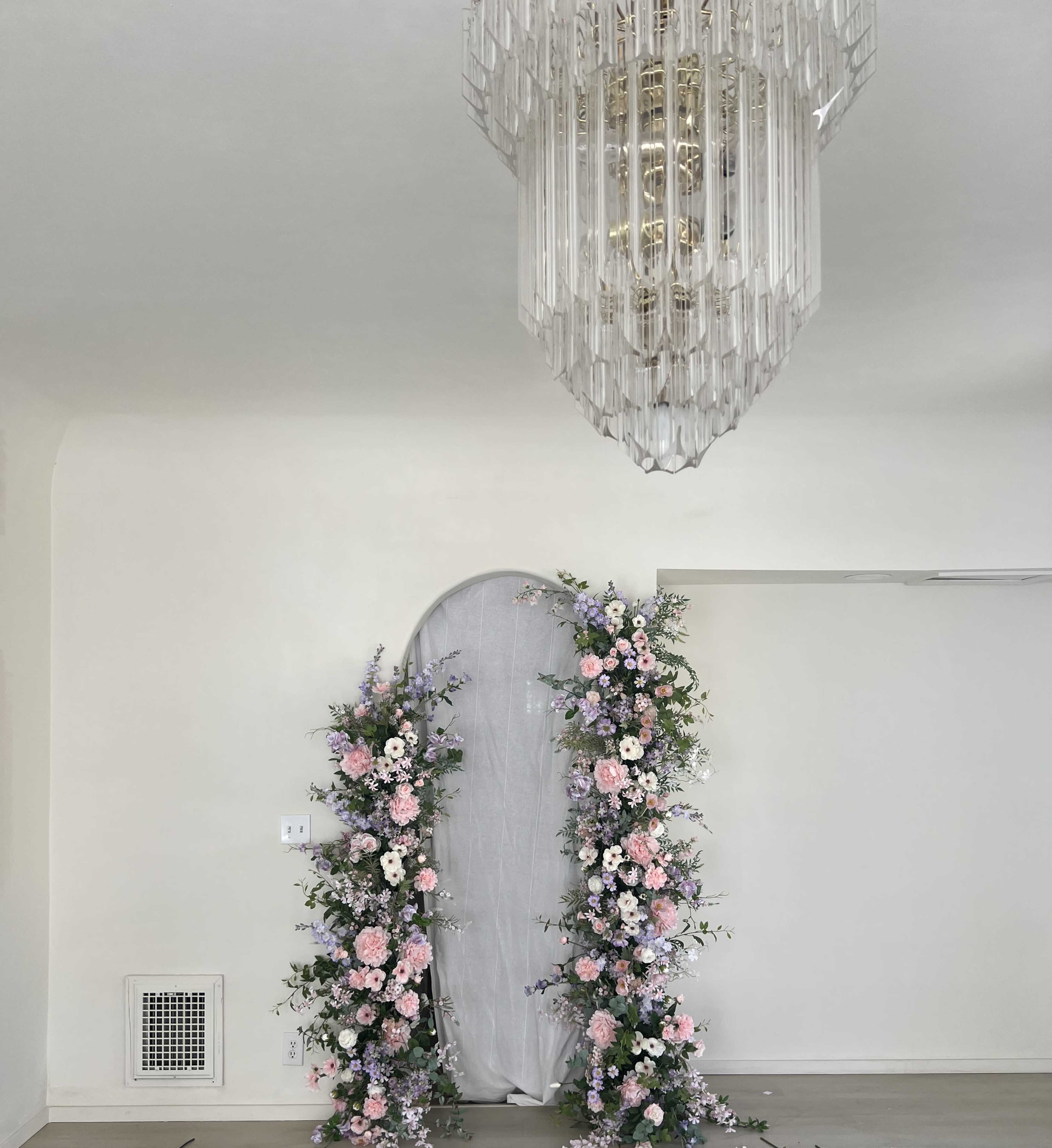 The image shows a floral arch adorned with pink and purple flowers in front of a light gray curtain, beneath a geometric crystal chandelier in a bright room.