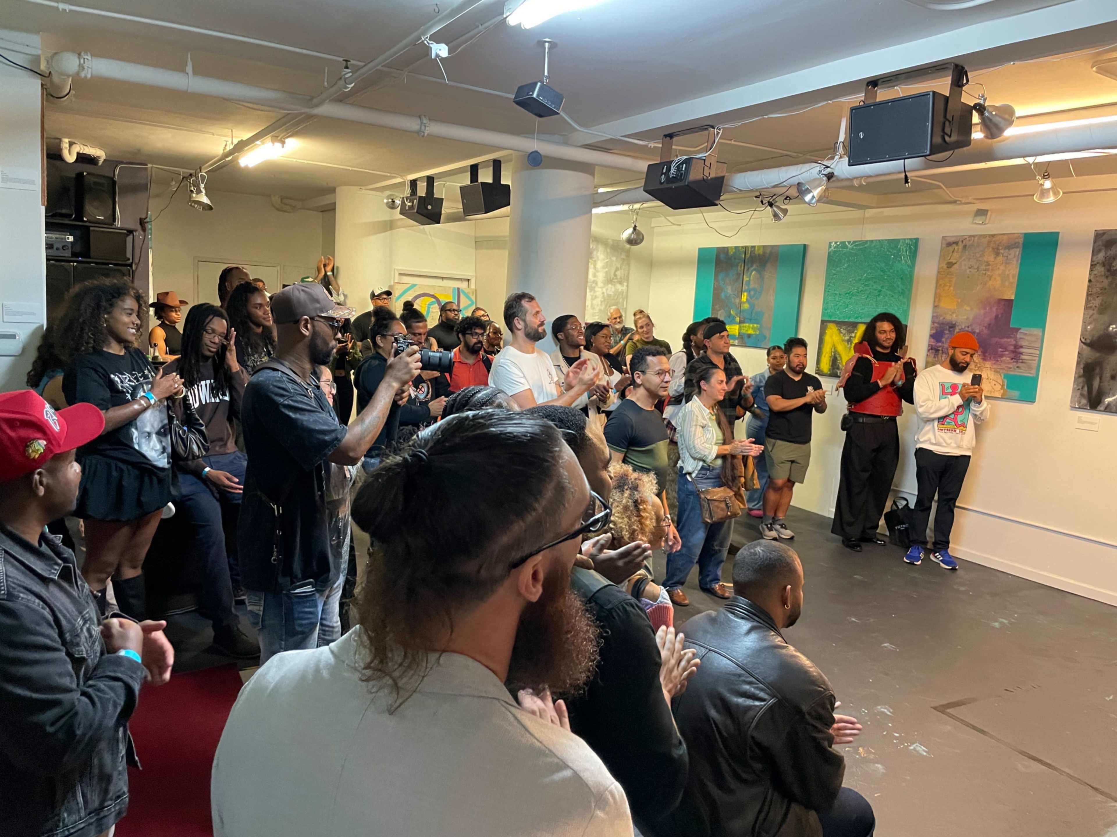 A crowd of people is gathered in an art gallery, clapping and engaging with the displayed artworks.