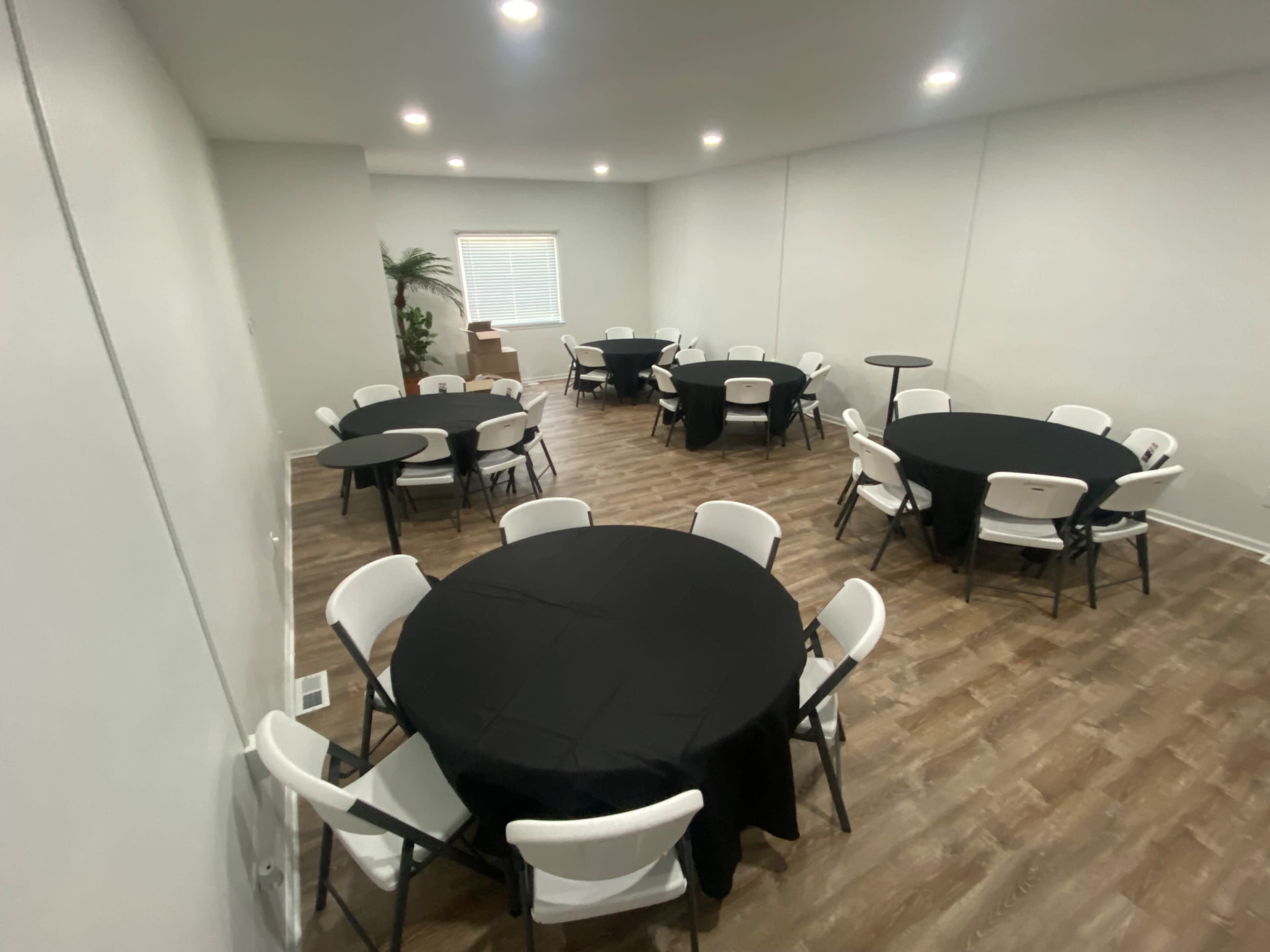 A well-lit room with several round tables covered in black tablecloths and arranged with white folding chairs around them.