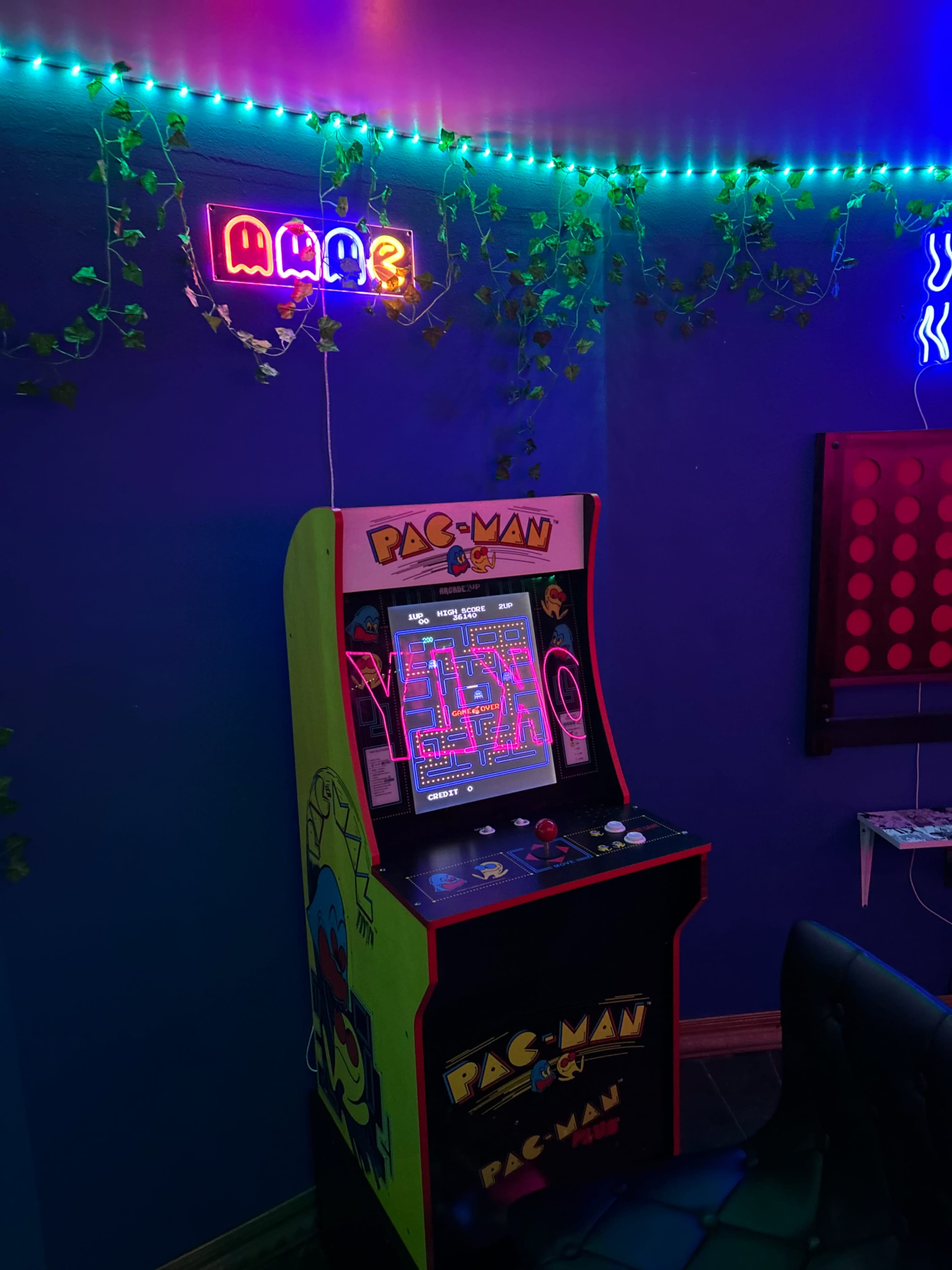 The image shows a vintage Pac-Man arcade machine in a room decorated with neon lights and greenery on the walls.