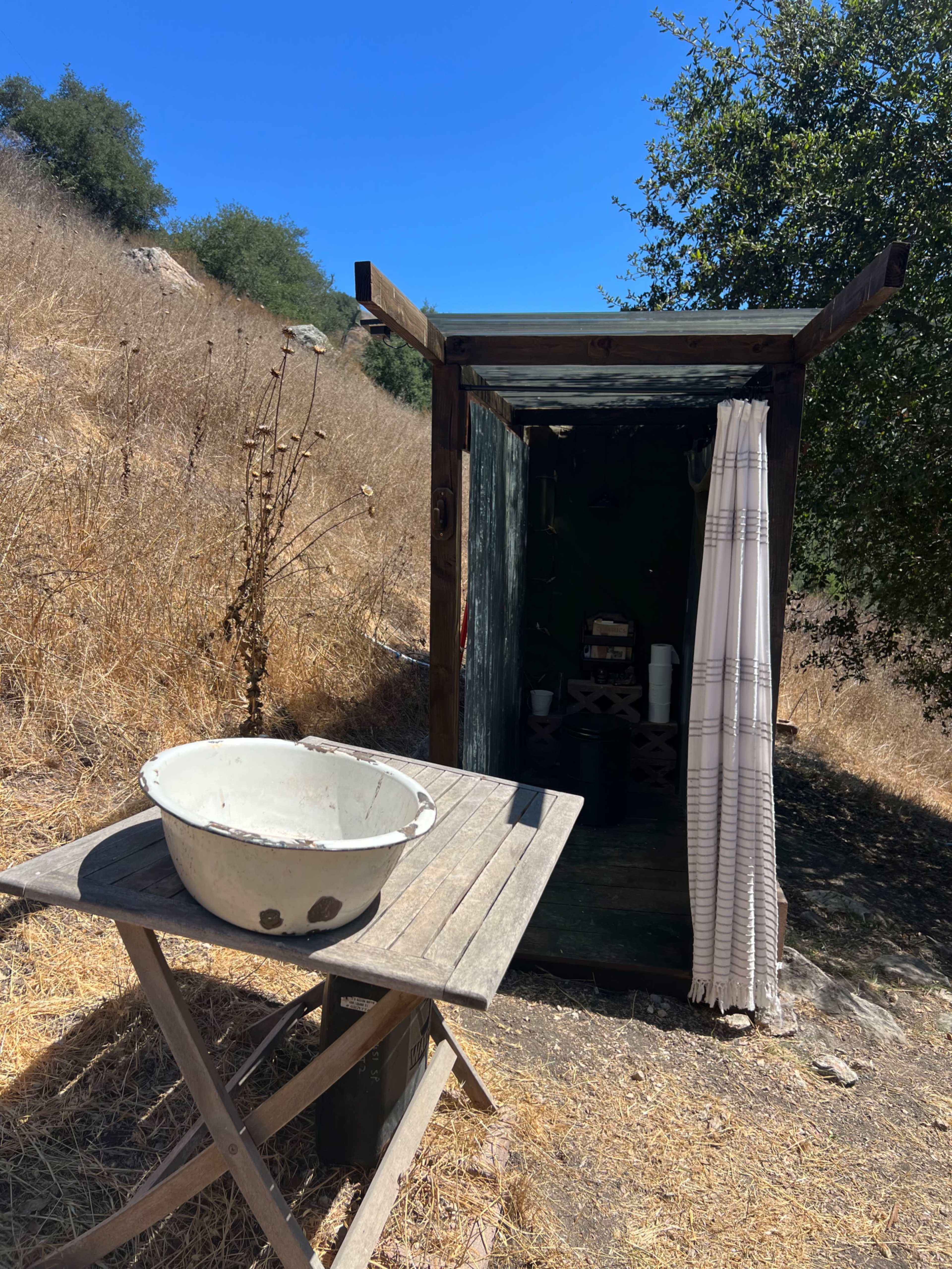 A small, rustic structure with a wooden exterior is nestled in a dry, grassy landscape, accompanied by a table holding a basin.