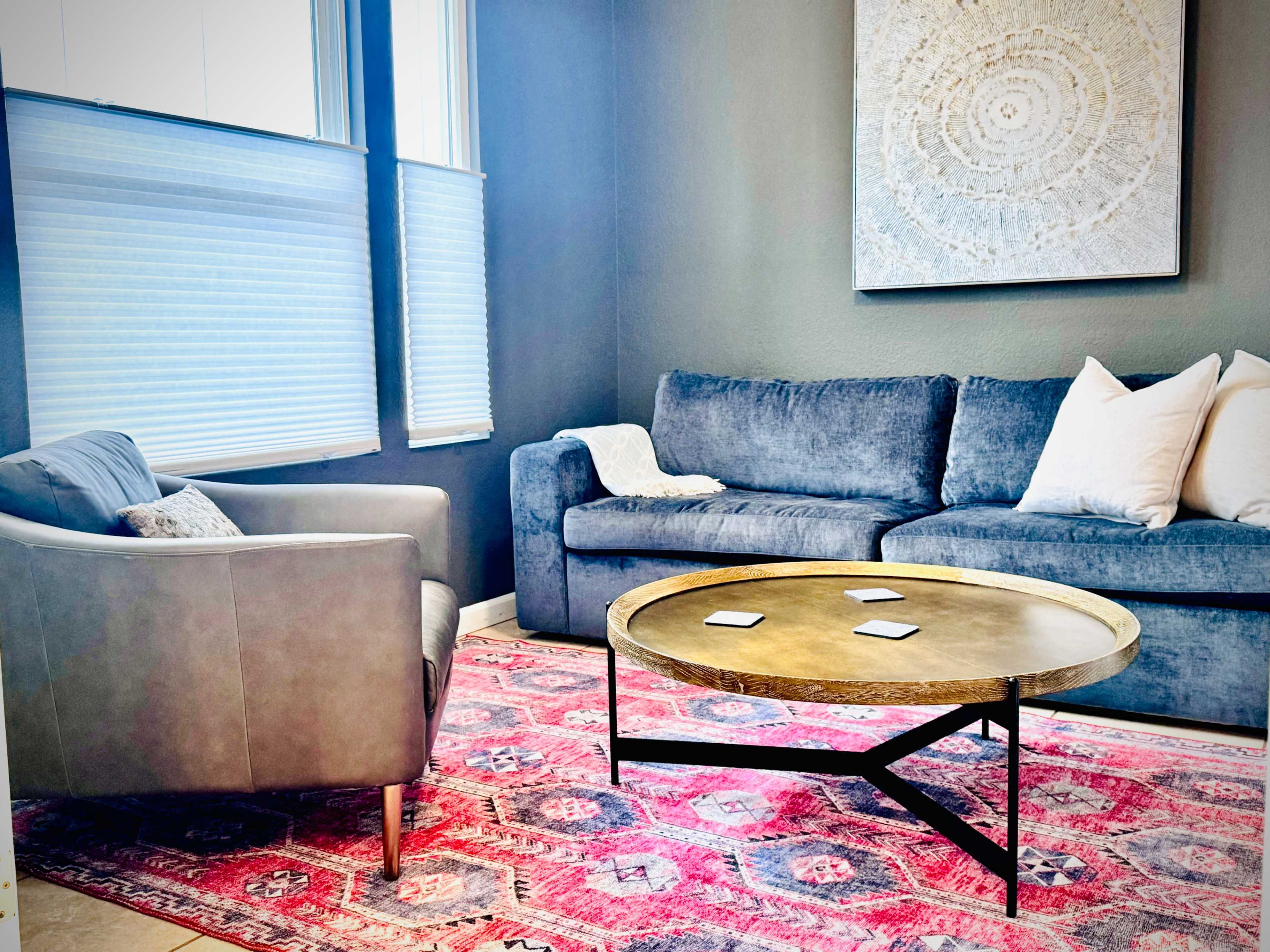 The image shows a cozy living room with a gray sofa, a light gray armchair, a round coffee table, and a vibrant area rug, with large windows allowing natural light to enter.