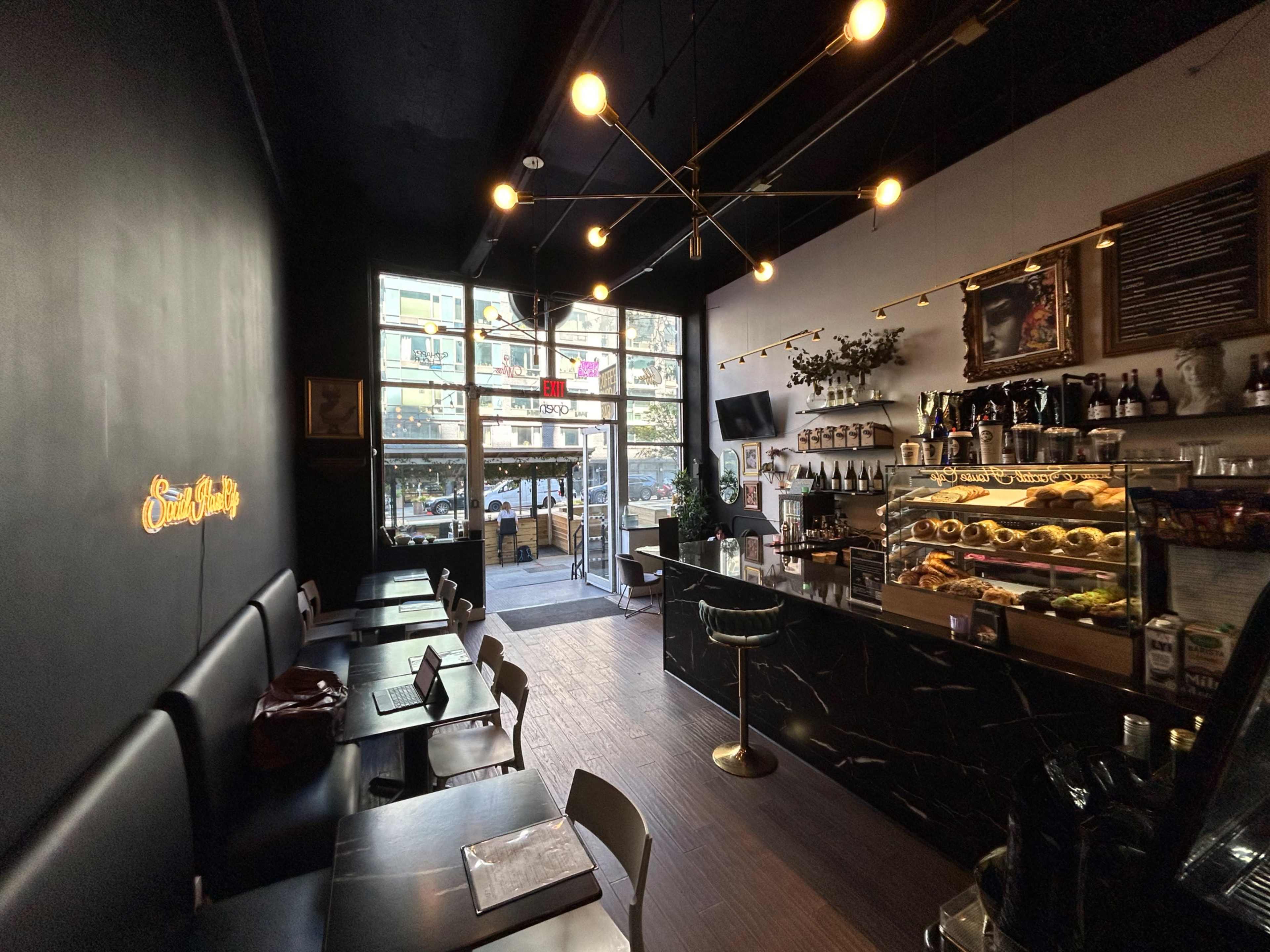 A modern café interior features dark seating, a glass front showcasing the street outside, and a display counter filled with pastries and beverages.