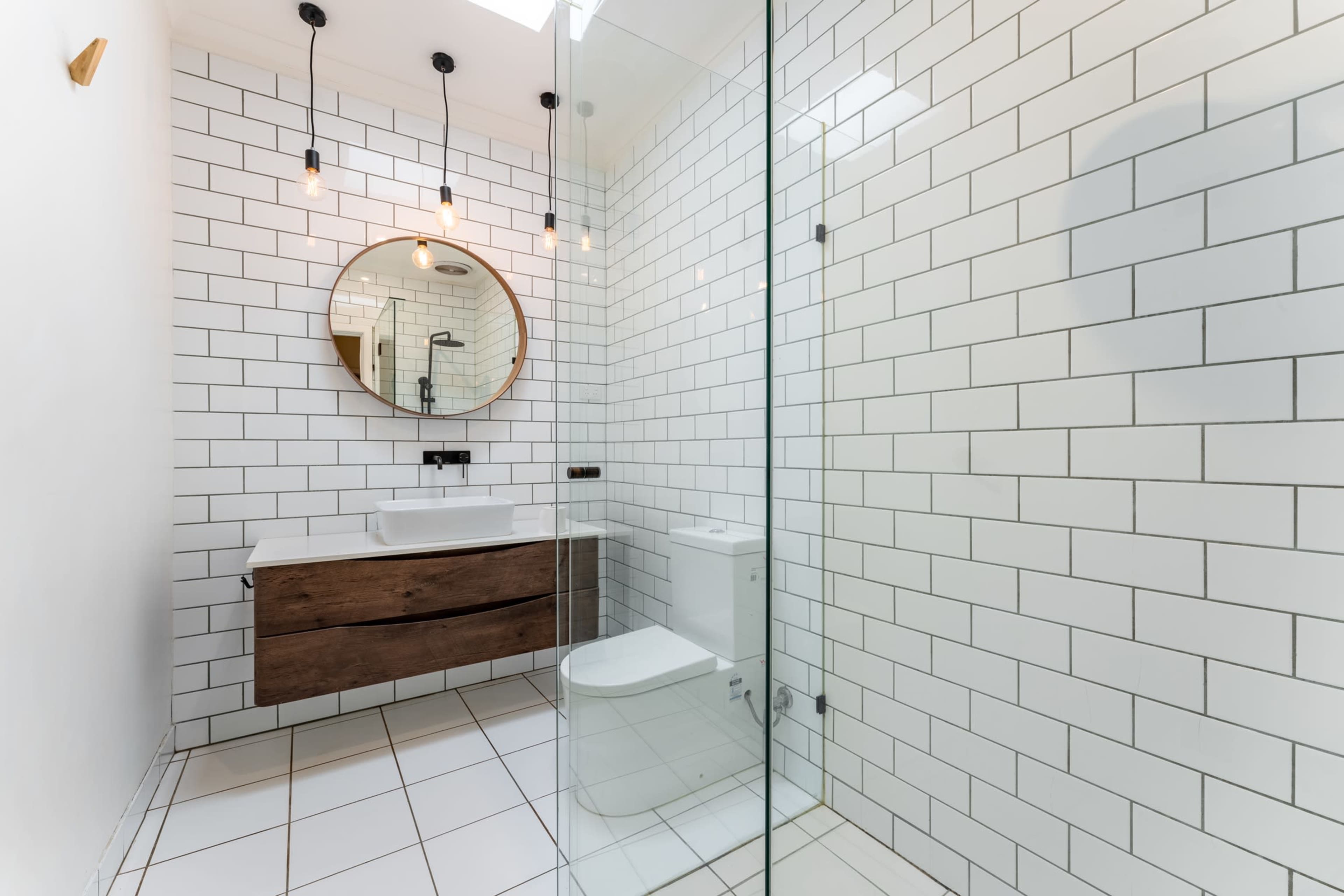 A modern bathroom featuring white subway tiles, a round mirror, a wooden sink cabinet, and a glass shower enclosure.
