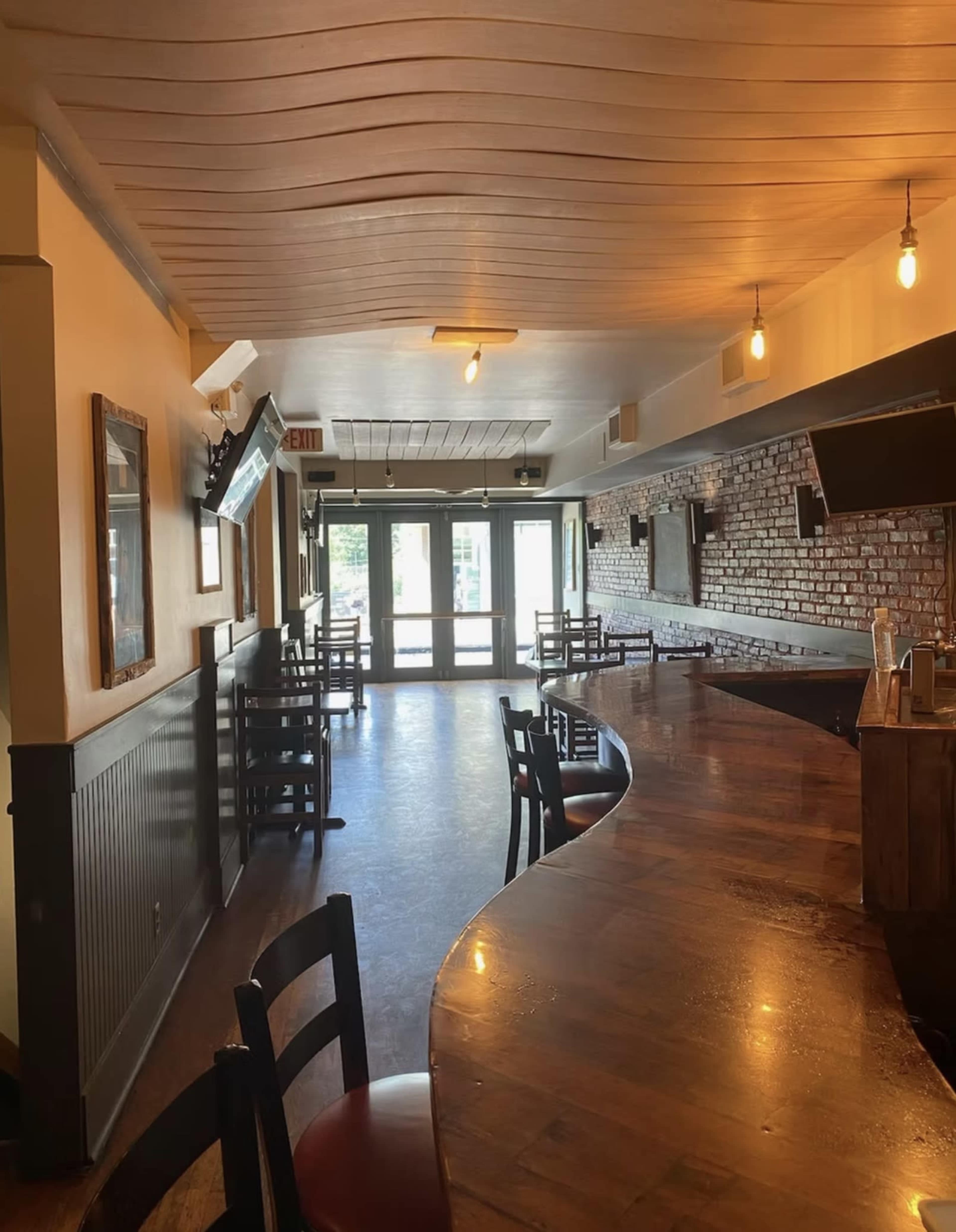The image shows a partially empty bar area with wooden furniture, exposed brick walls, and large windows allowing natural light to enter.