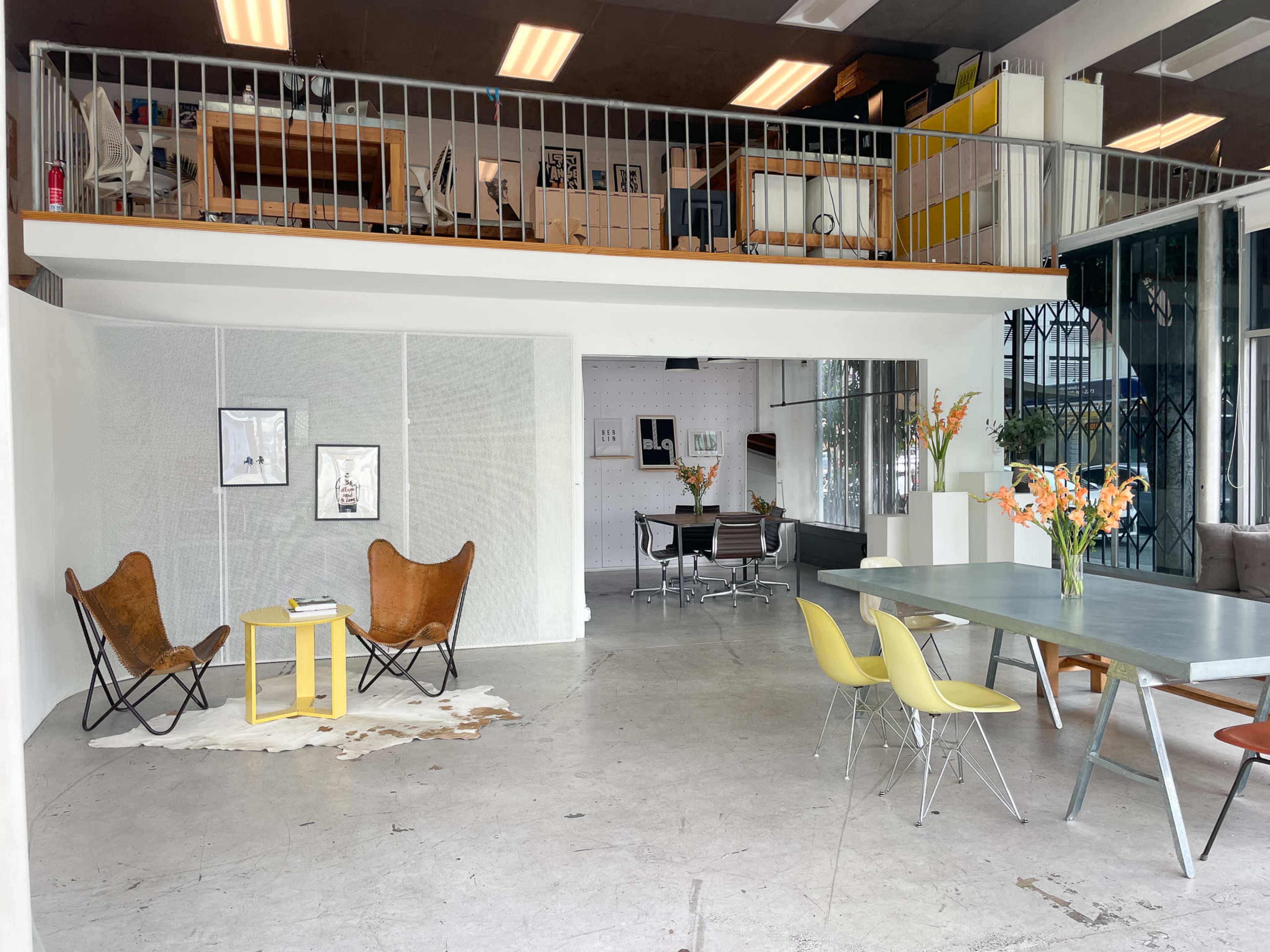 The image shows a spacious, modern office interior featuring a seating area with two butterfly chairs and a yellow table, alongside a dining table and chairs, with a lofted workspace visible above.