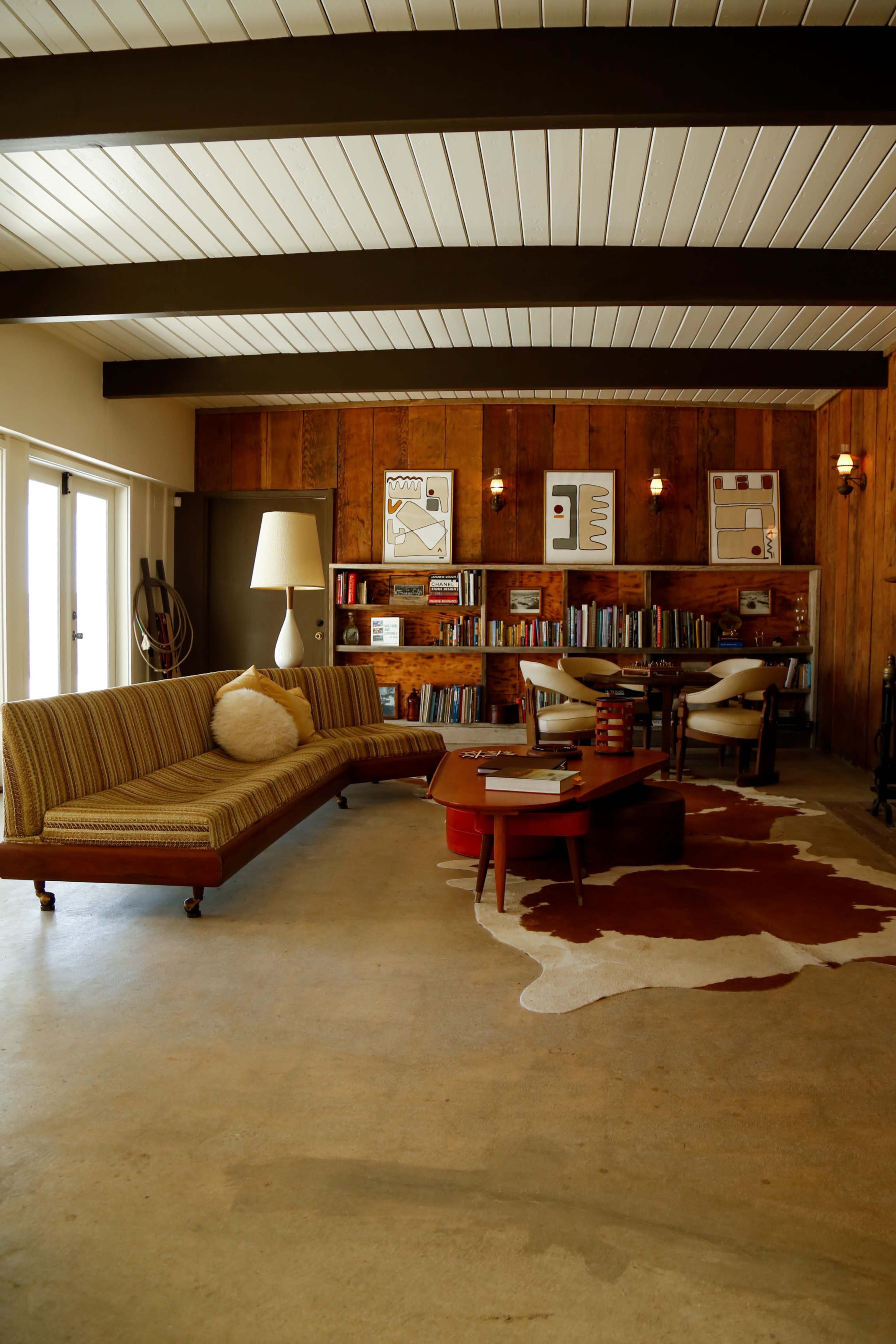 A mid-century modern living room featuring a striped sofa, a wooden bookshelf filled with books, and a cowhide rug on a polished concrete floor.