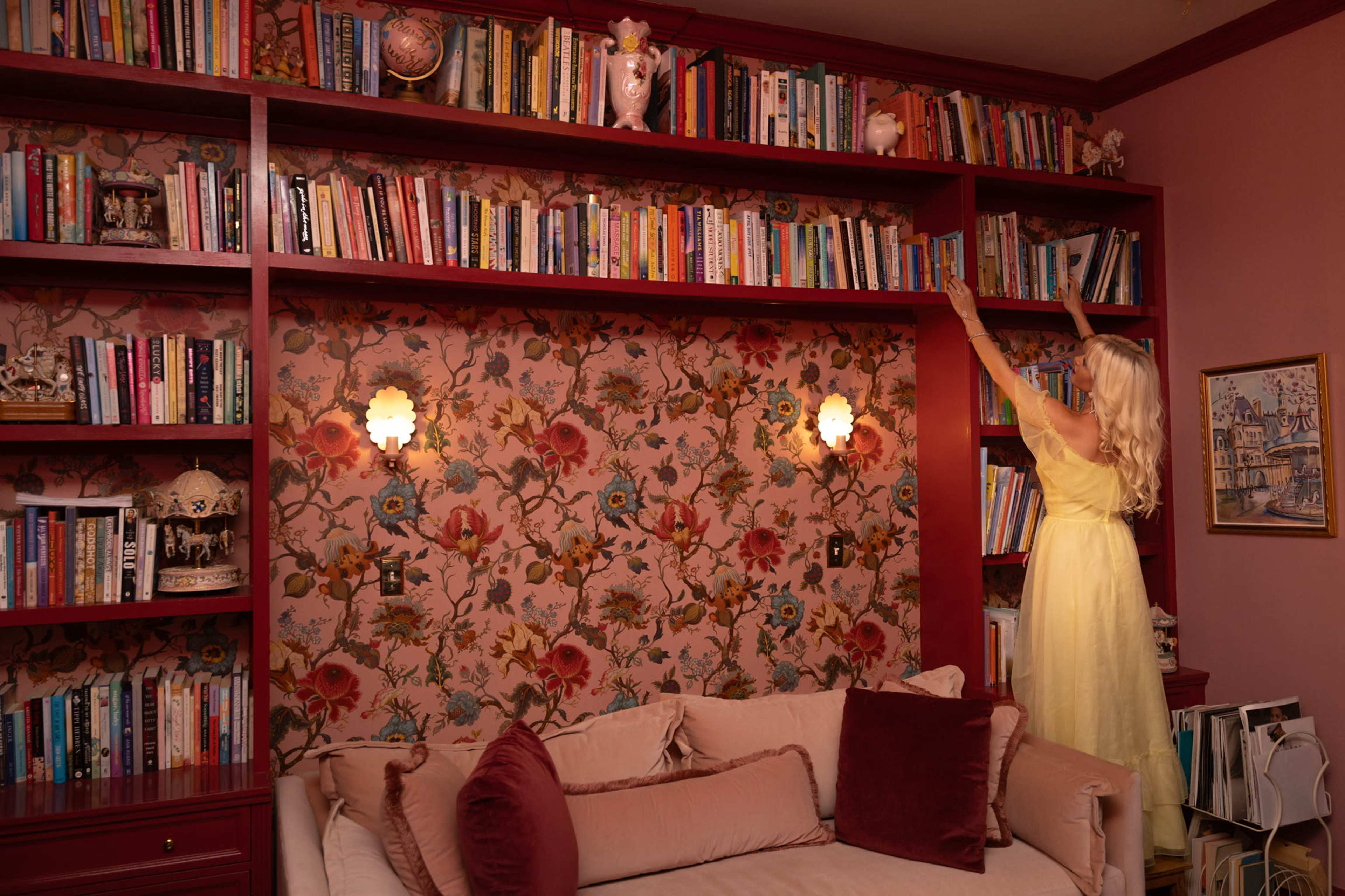A woman in a yellow dress reaches for a book on a shelf in a room with floral wallpaper and a plush sofa.