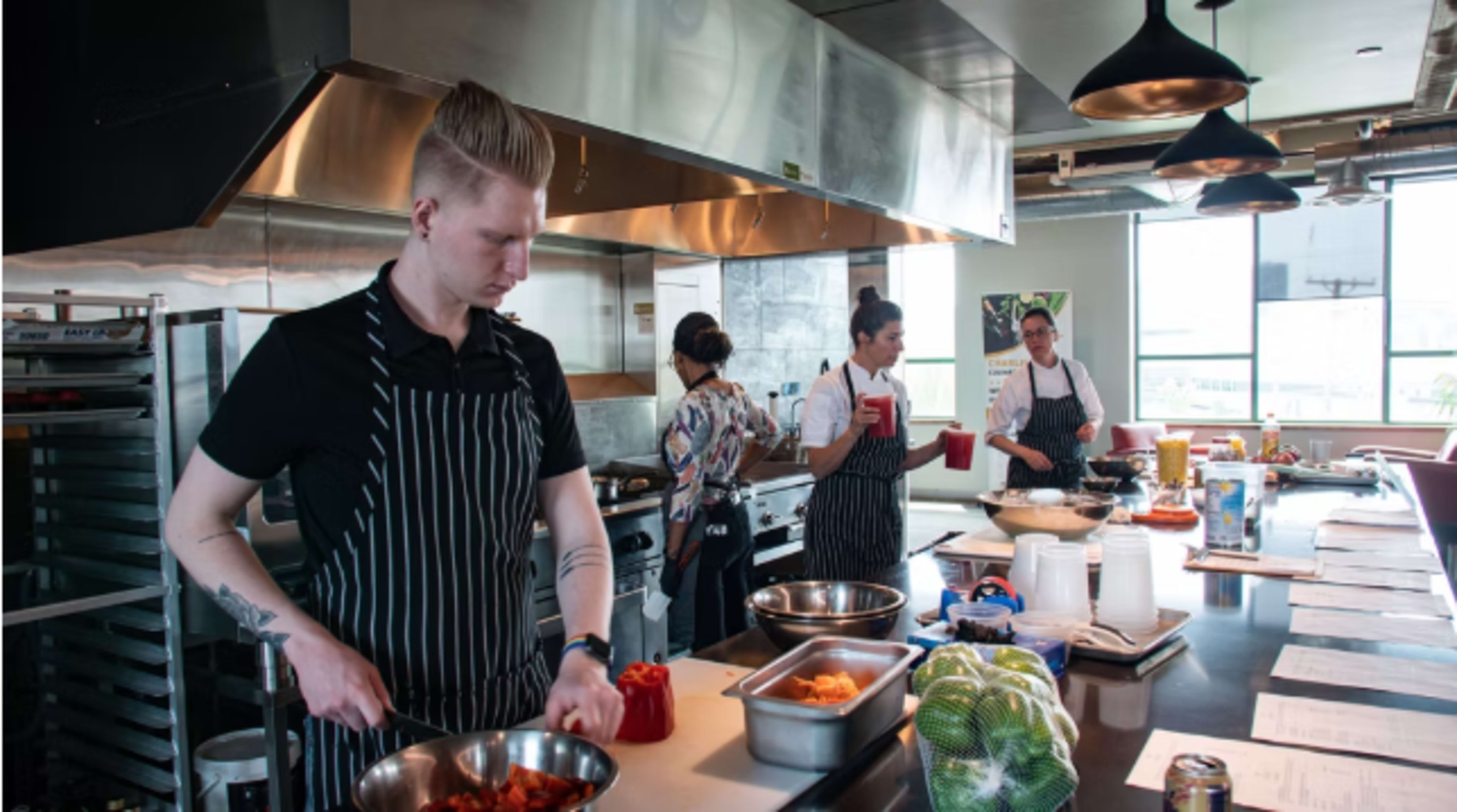 Three chefs are preparing ingredients in a modern kitchen with stainless steel appliances, while one chef chops vegetables at a counter.