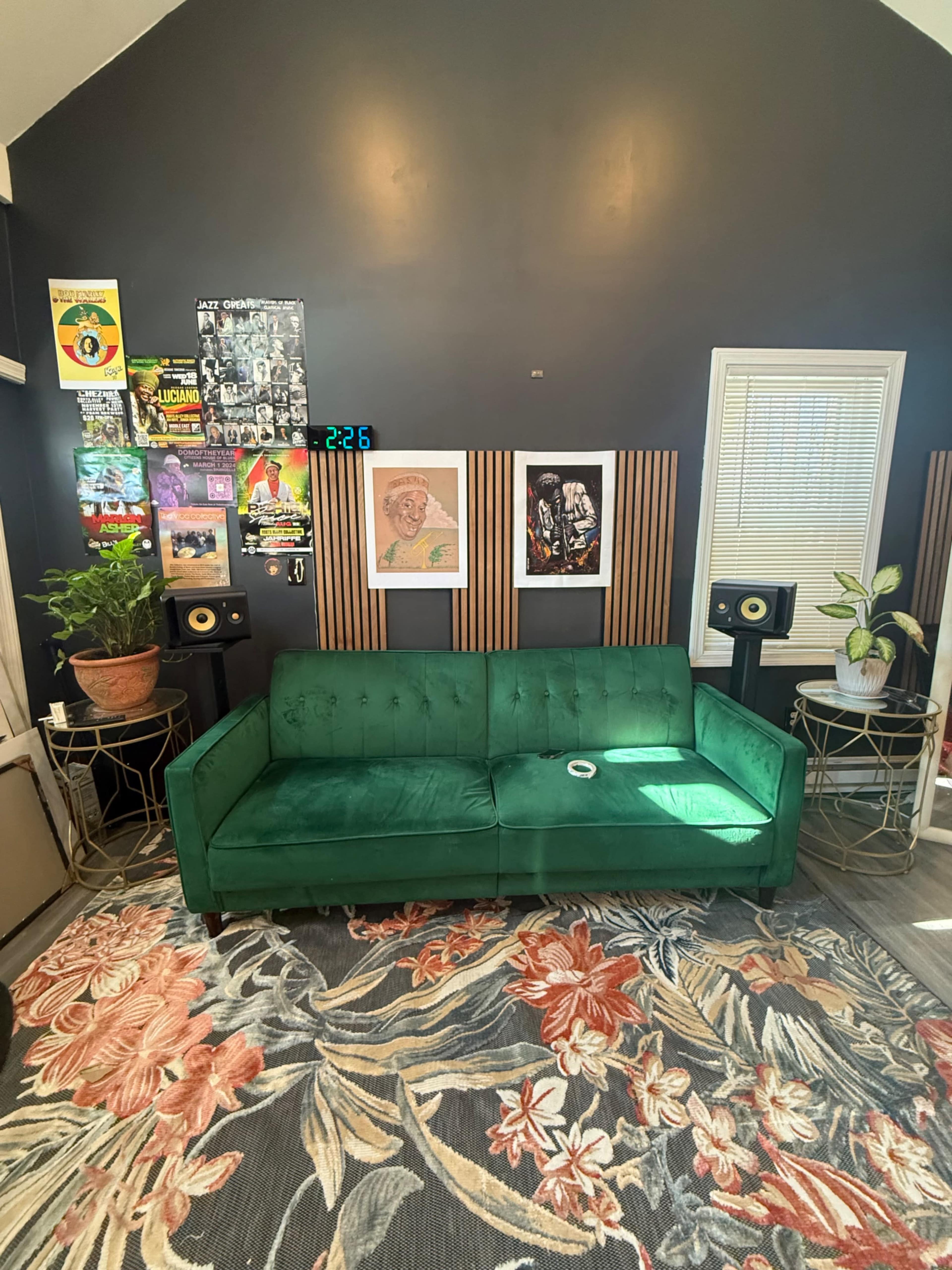 The image shows a green velvet sofa set against a dark wall decorated with various framed artworks and a clock, alongside two small tables with speakers and potted plants.