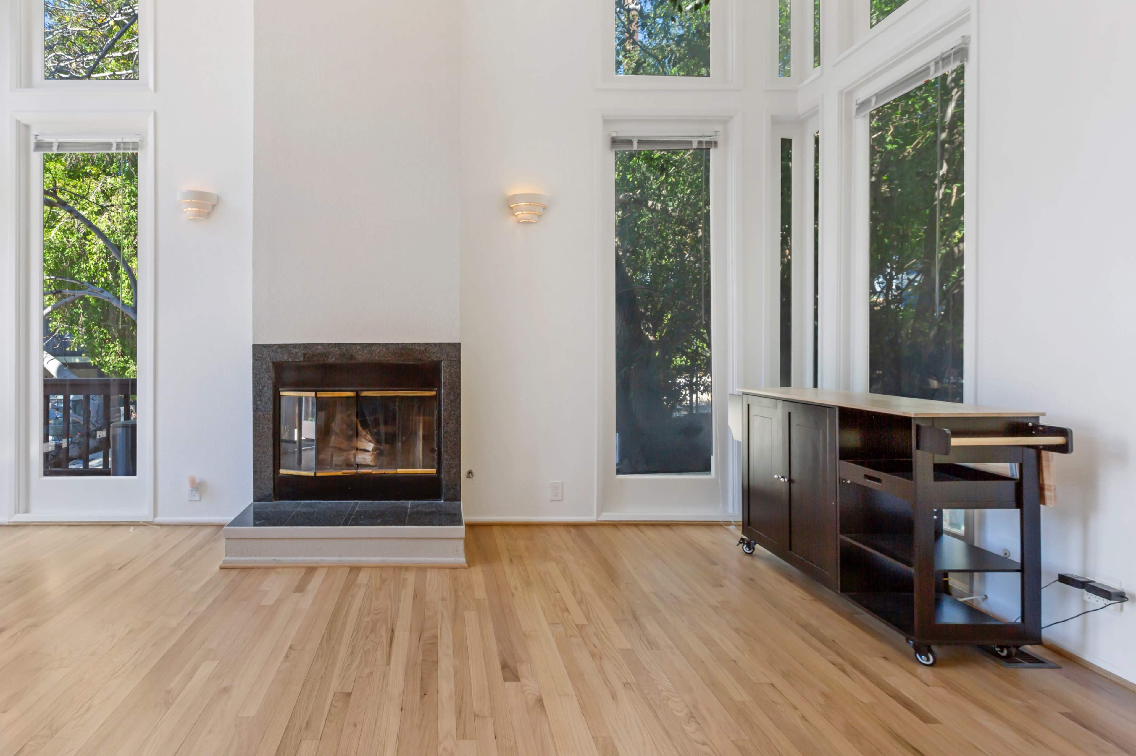 The image shows a bright living room with large windows, a modern fireplace, and a wooden cabinet on wheels.
