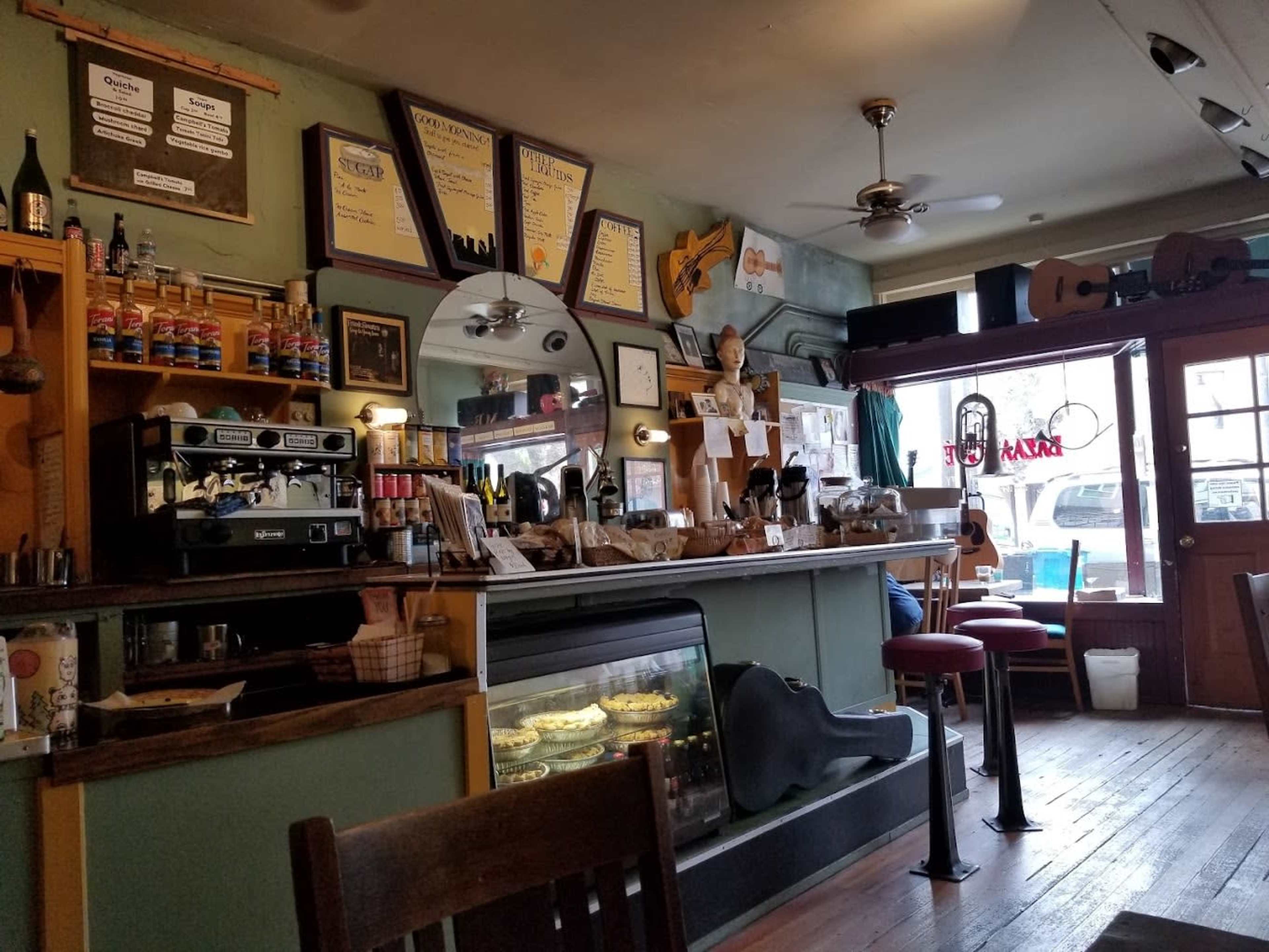 The interior of a café features a counter with an espresso machine, baked goods, musical instruments, and a seating area with wooden chairs and red stools.