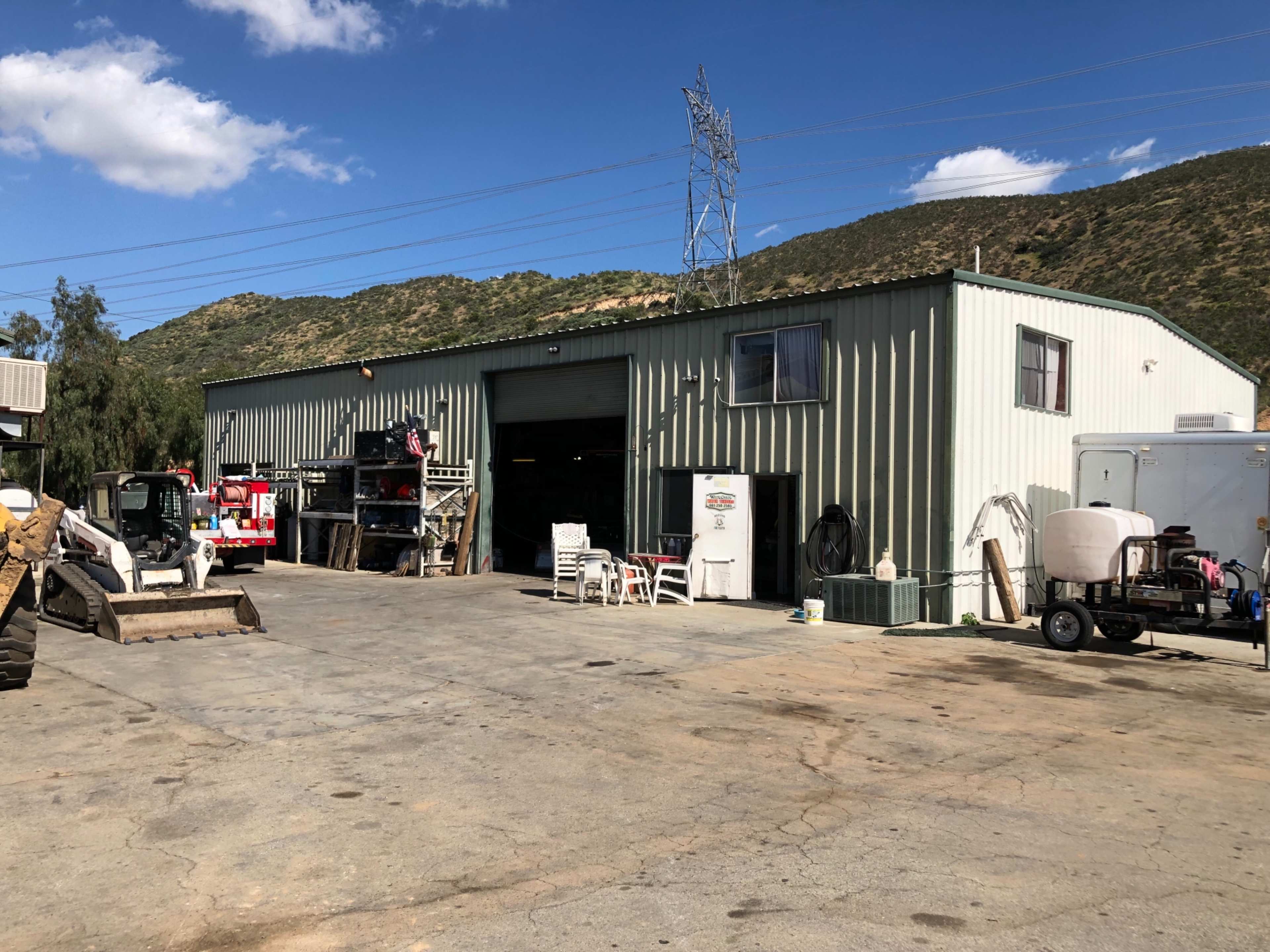 A metal building with an open garage door stands in an outdoor area surrounded by machinery and equipment.