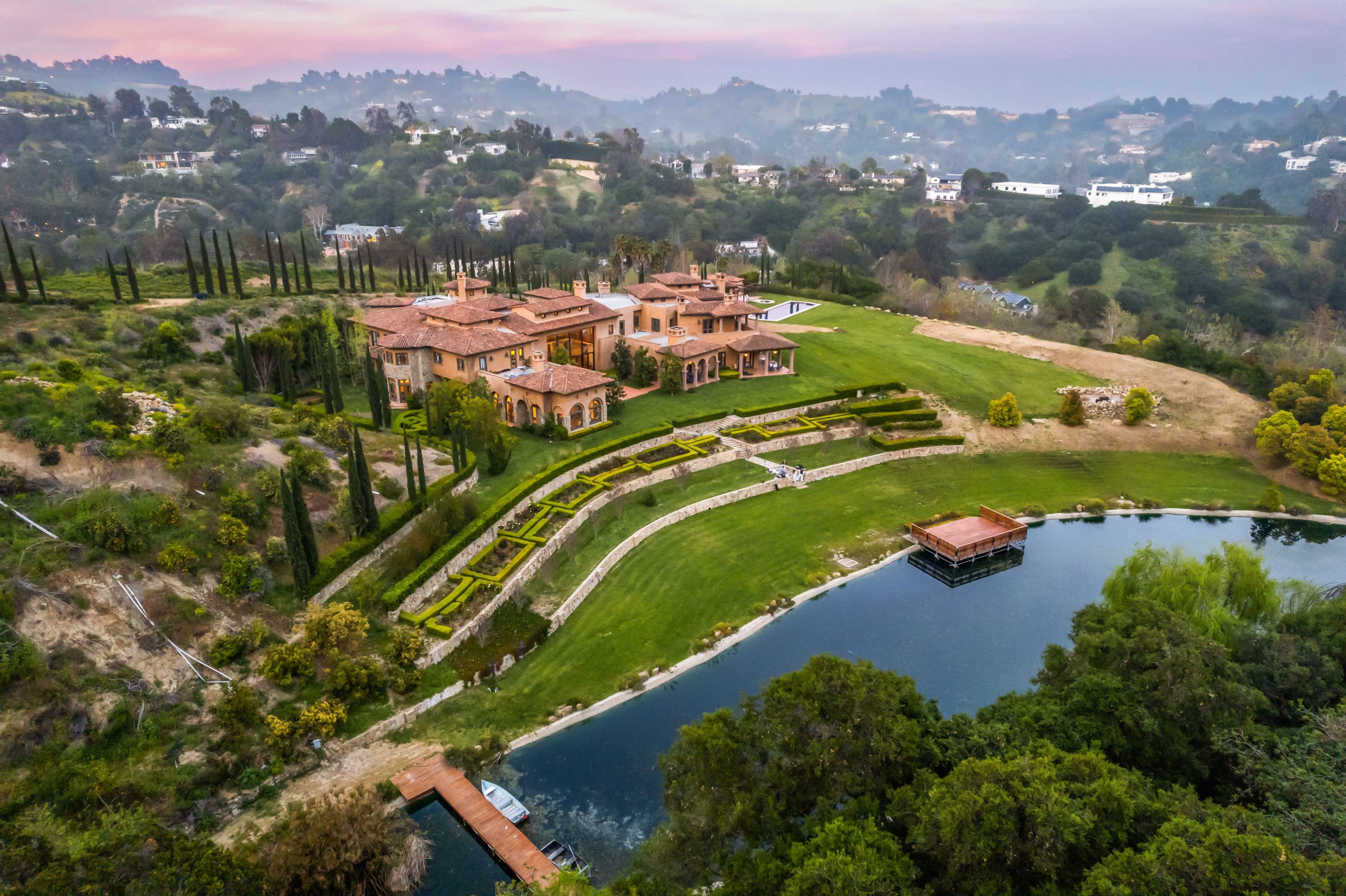 The image shows a sprawling estate with a large mansion, manicured gardens, and a pond surrounded by lush hills and trees.