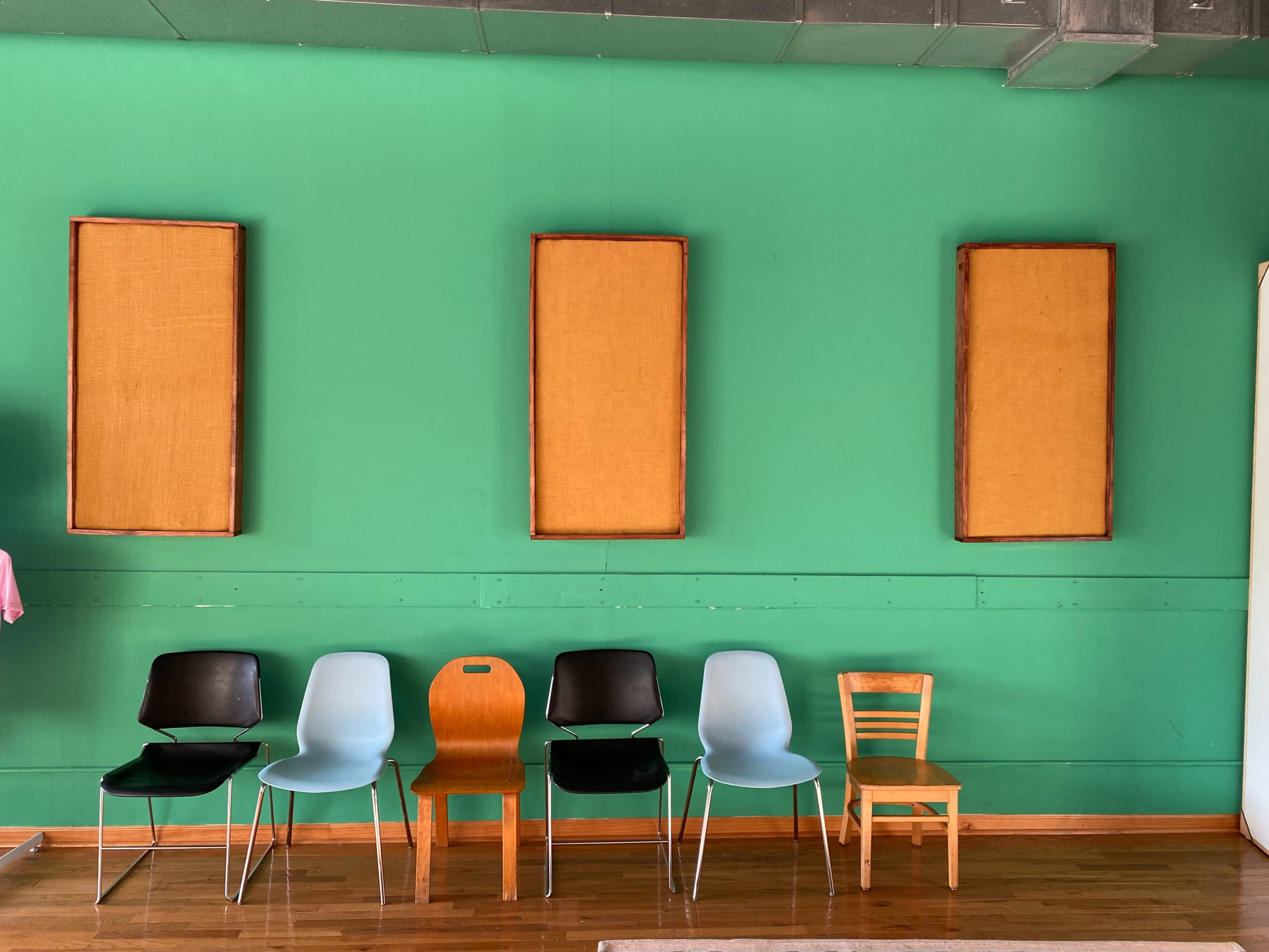A row of six chairs of different styles and colors is lined up against a green wall, with three empty frames hanging above them.