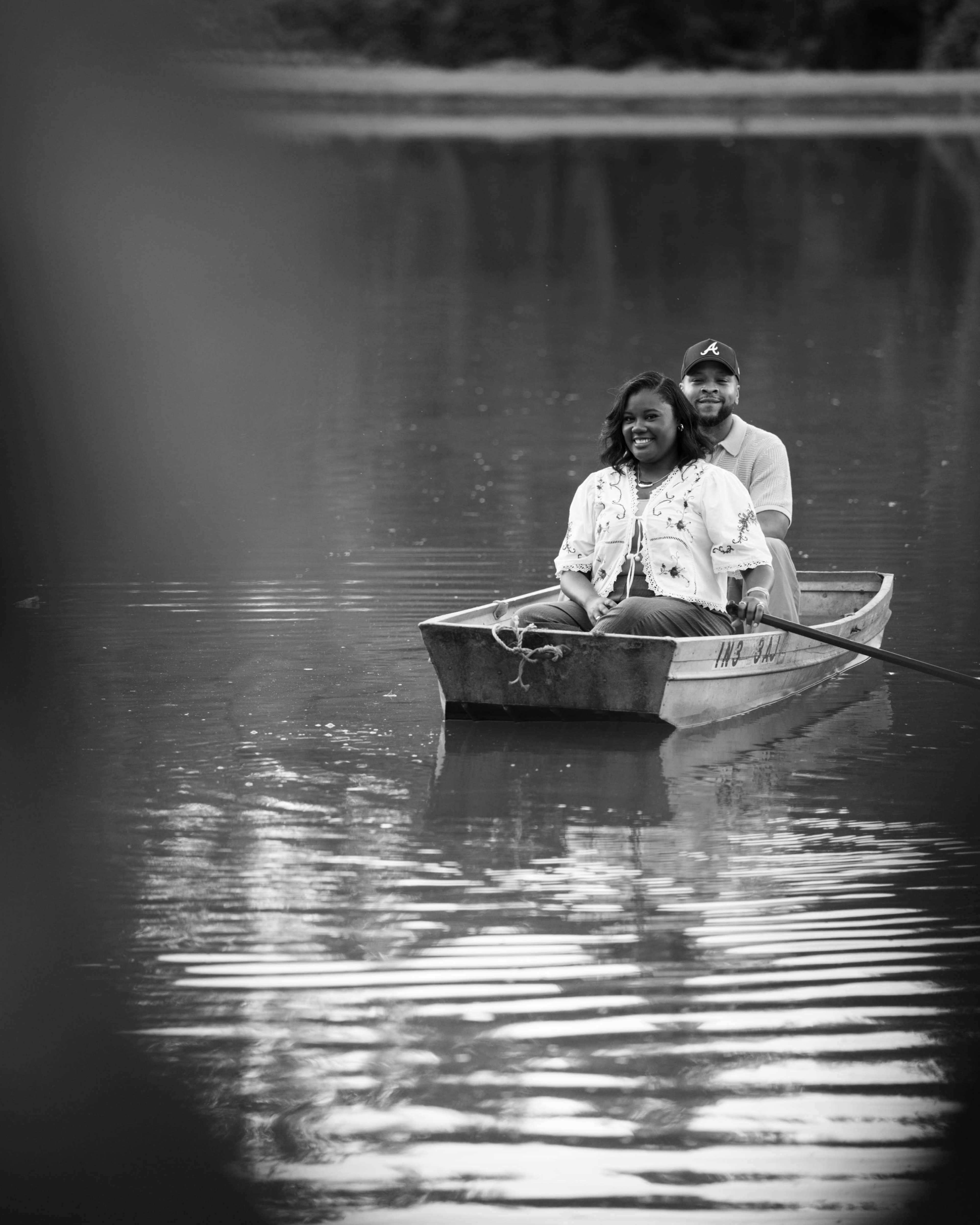 A couple rows a small boat on a calm water surface surrounded by trees.