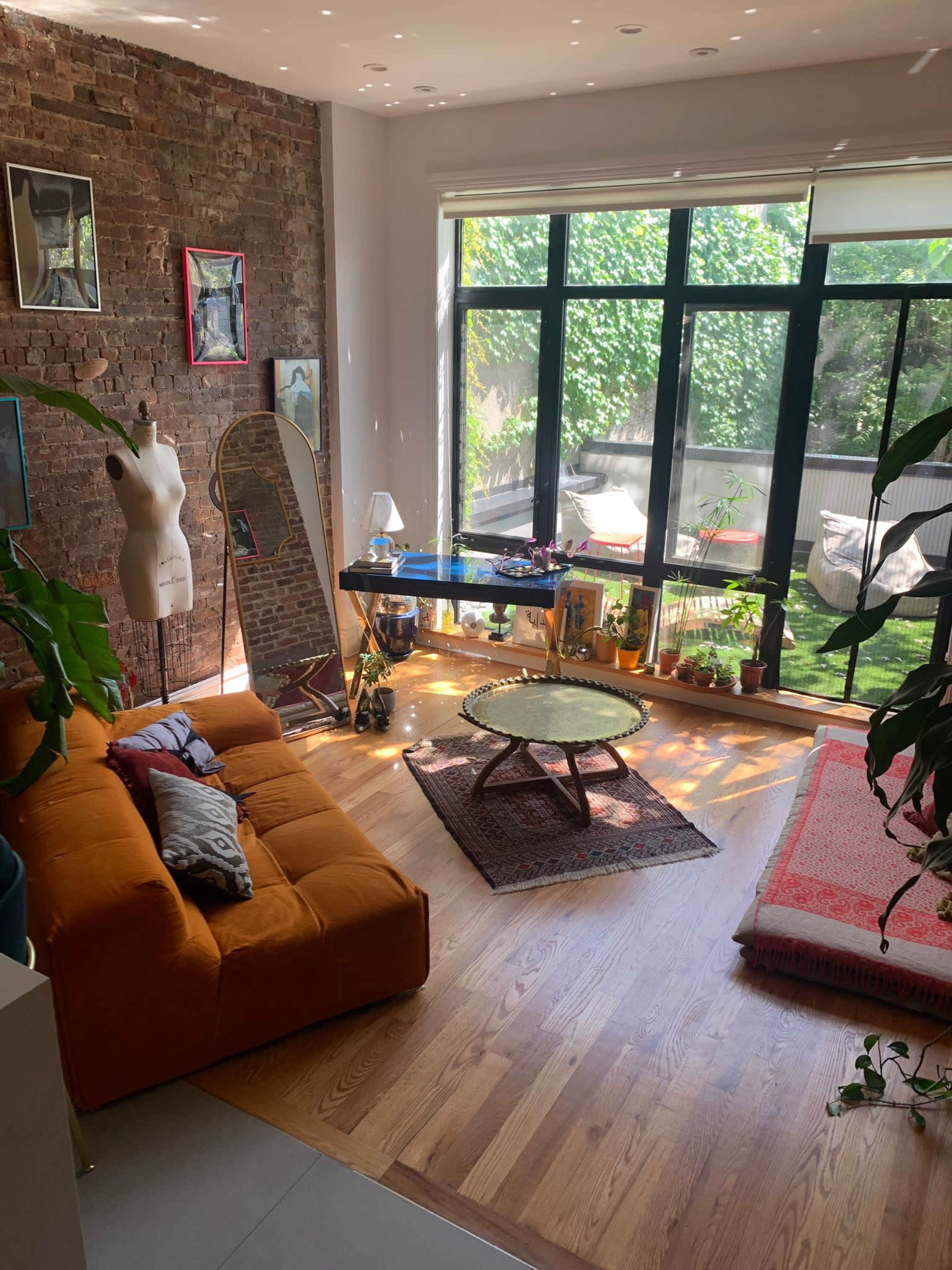 The image shows a bright living room featuring a brick wall, wooden flooring, a yellow couch, a mirror, and large windows overlooking greenery.