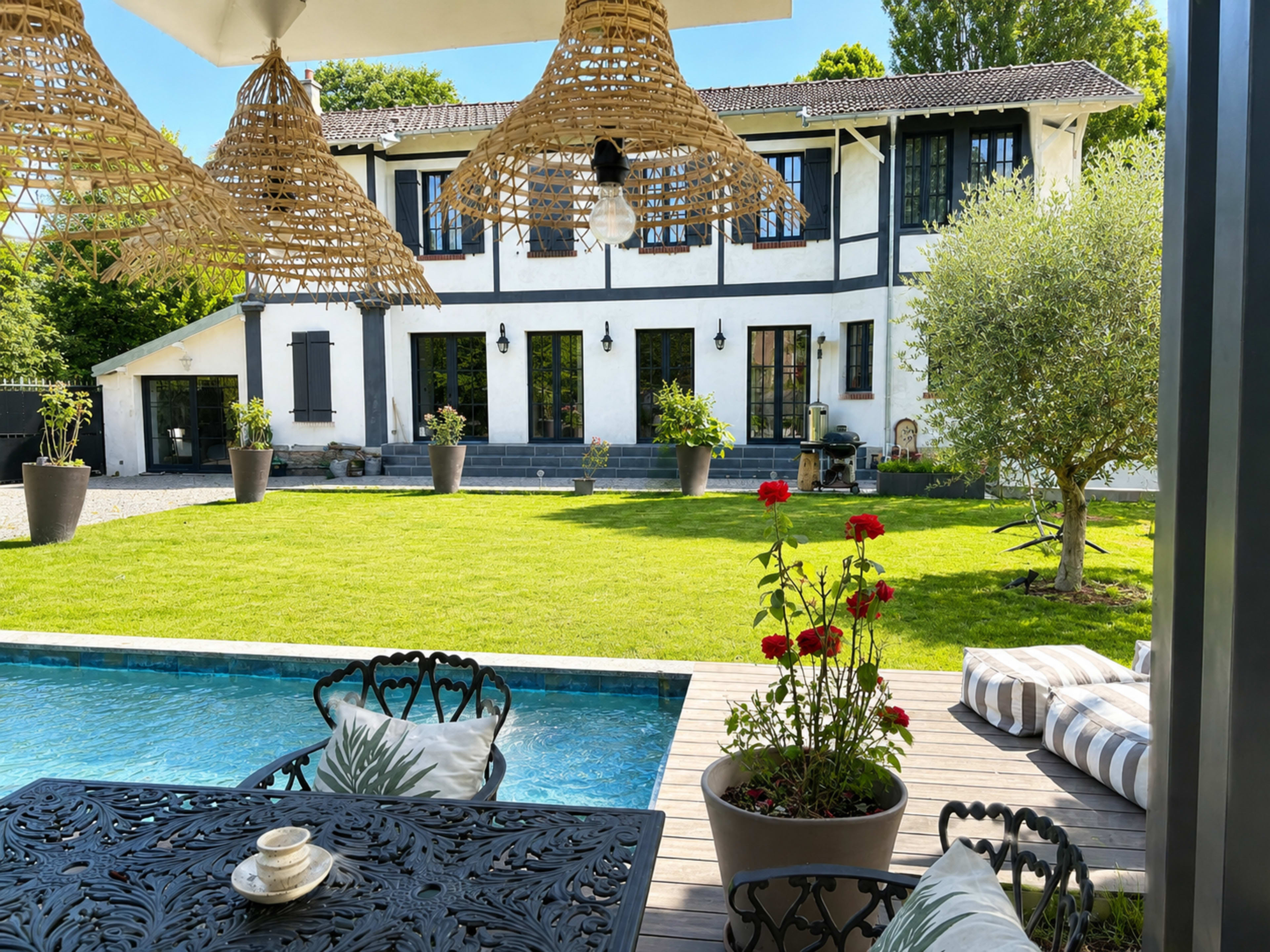 The image shows a well-maintained garden with a swimming pool and a stylish outdoor dining area in front of a two-story white house with black accents.