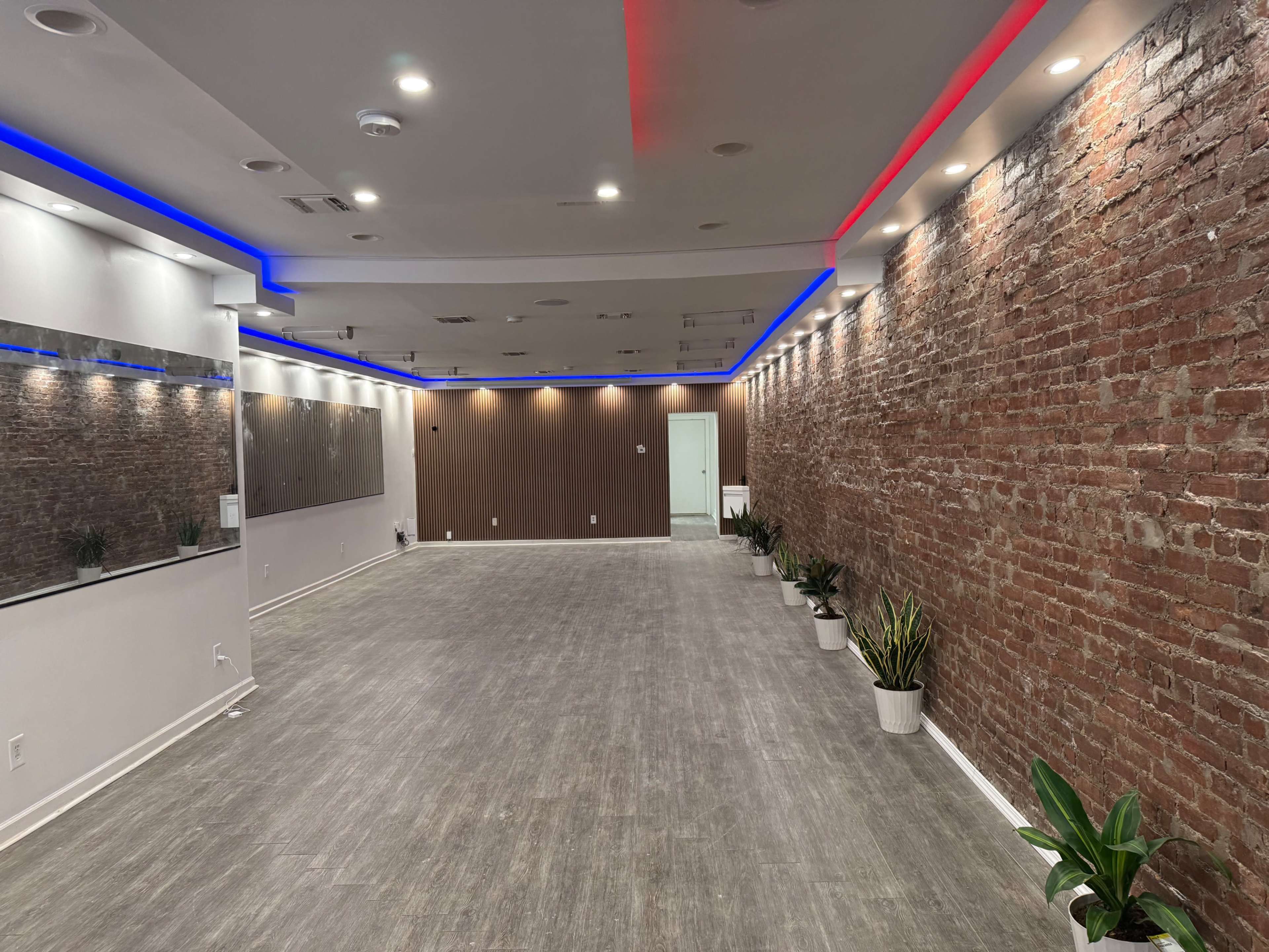 The image shows a spacious room with exposed brick walls, gray flooring, and modern lighting along the ceiling, accompanied by potted plants.