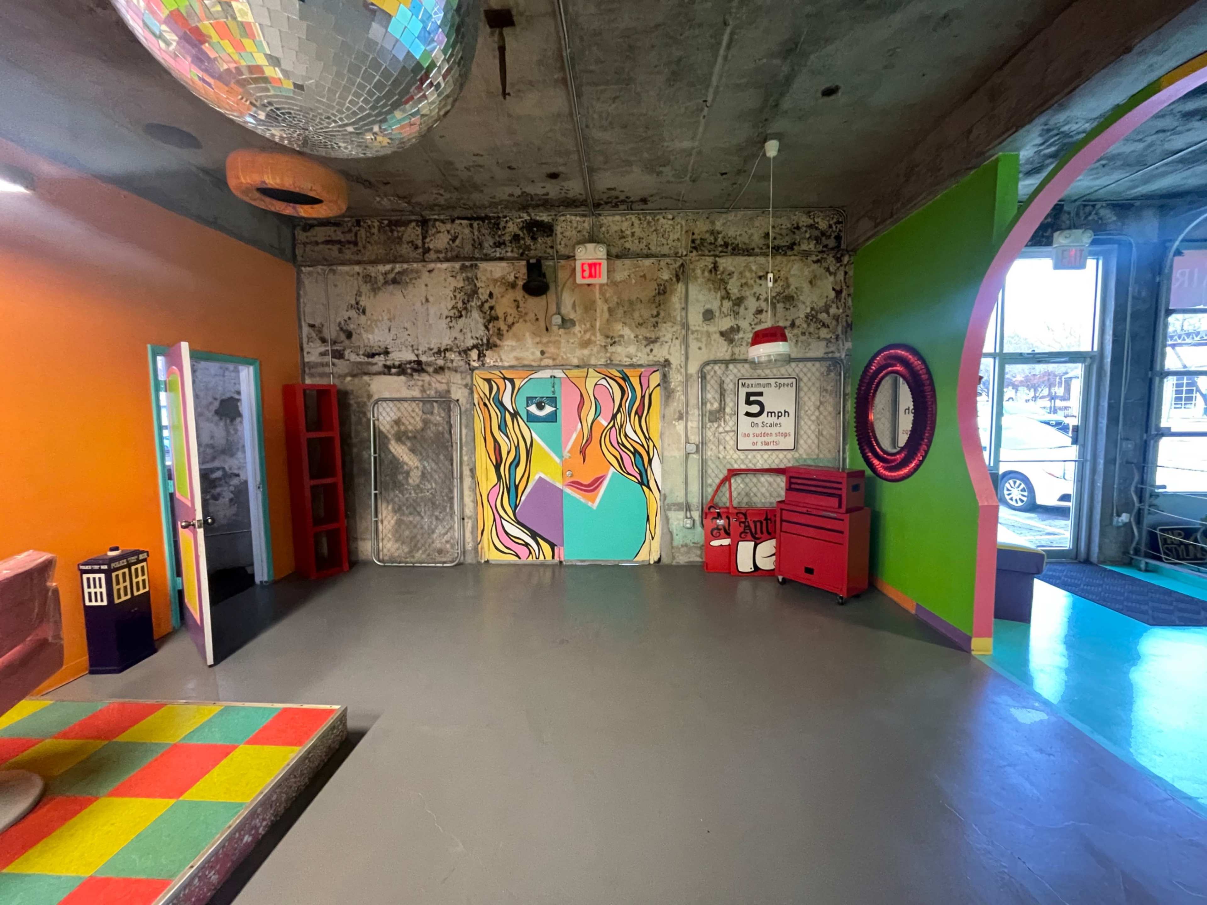 A colorful interior space features a mural of a face, a disco ball hanging from the ceiling, and various furniture items, including a red cart and patterned floor tiles.