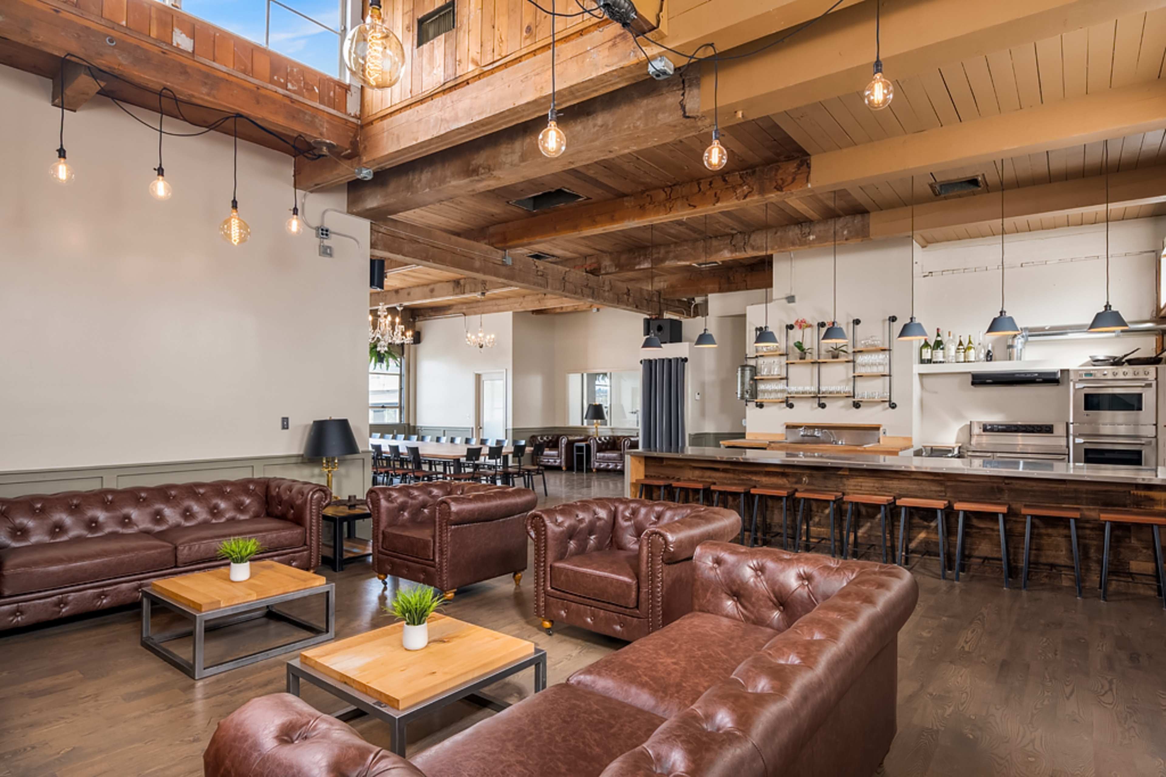 A spacious, modern interior with leather couches arranged around wooden coffee tables and a kitchen area in the background featuring stainless steel appliances.