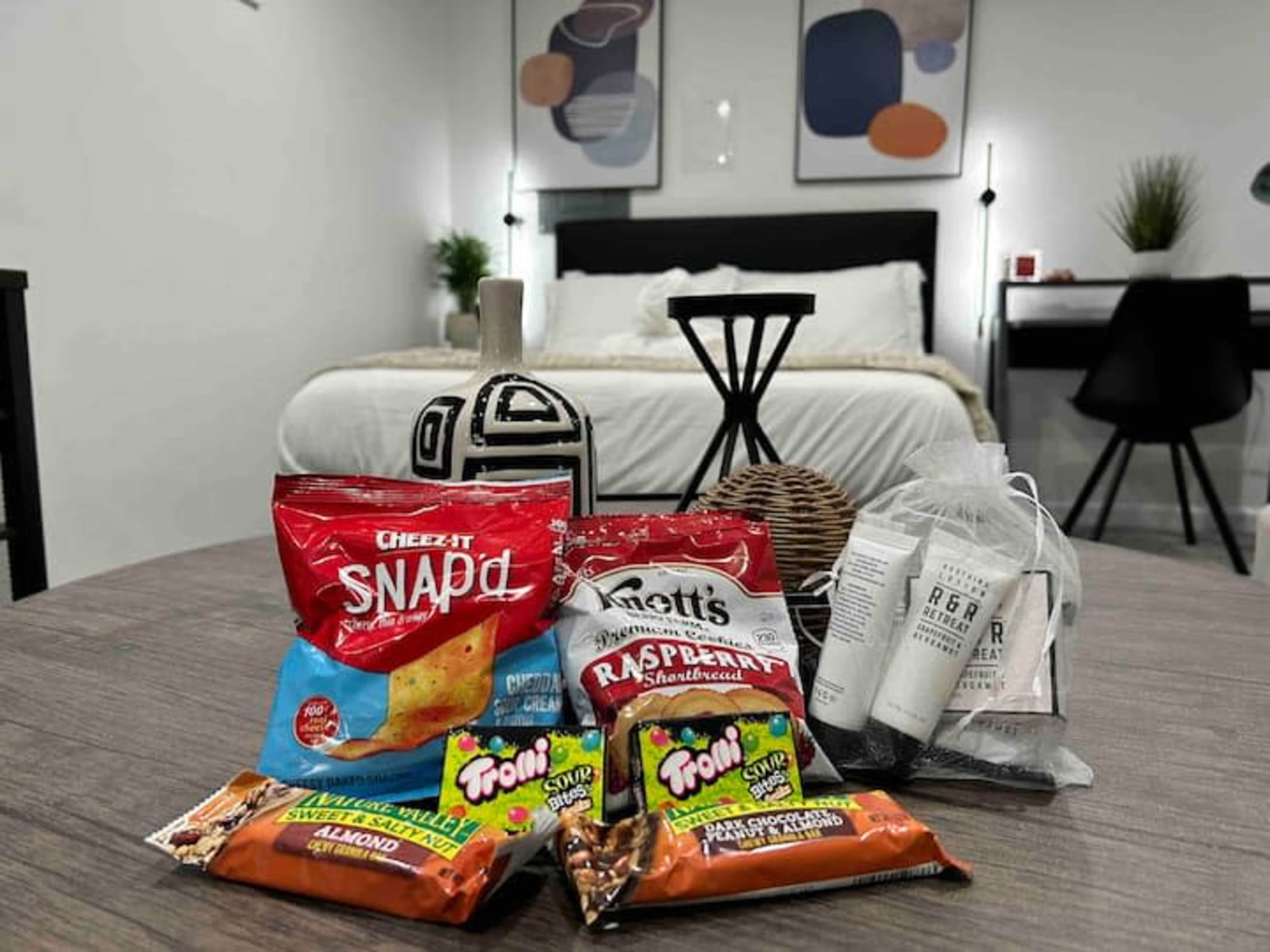A table with various snacks, including Cheez-It Snap'd and Knott's raspberry shortbread cookies, in a room featuring a bed and a desk in the background.