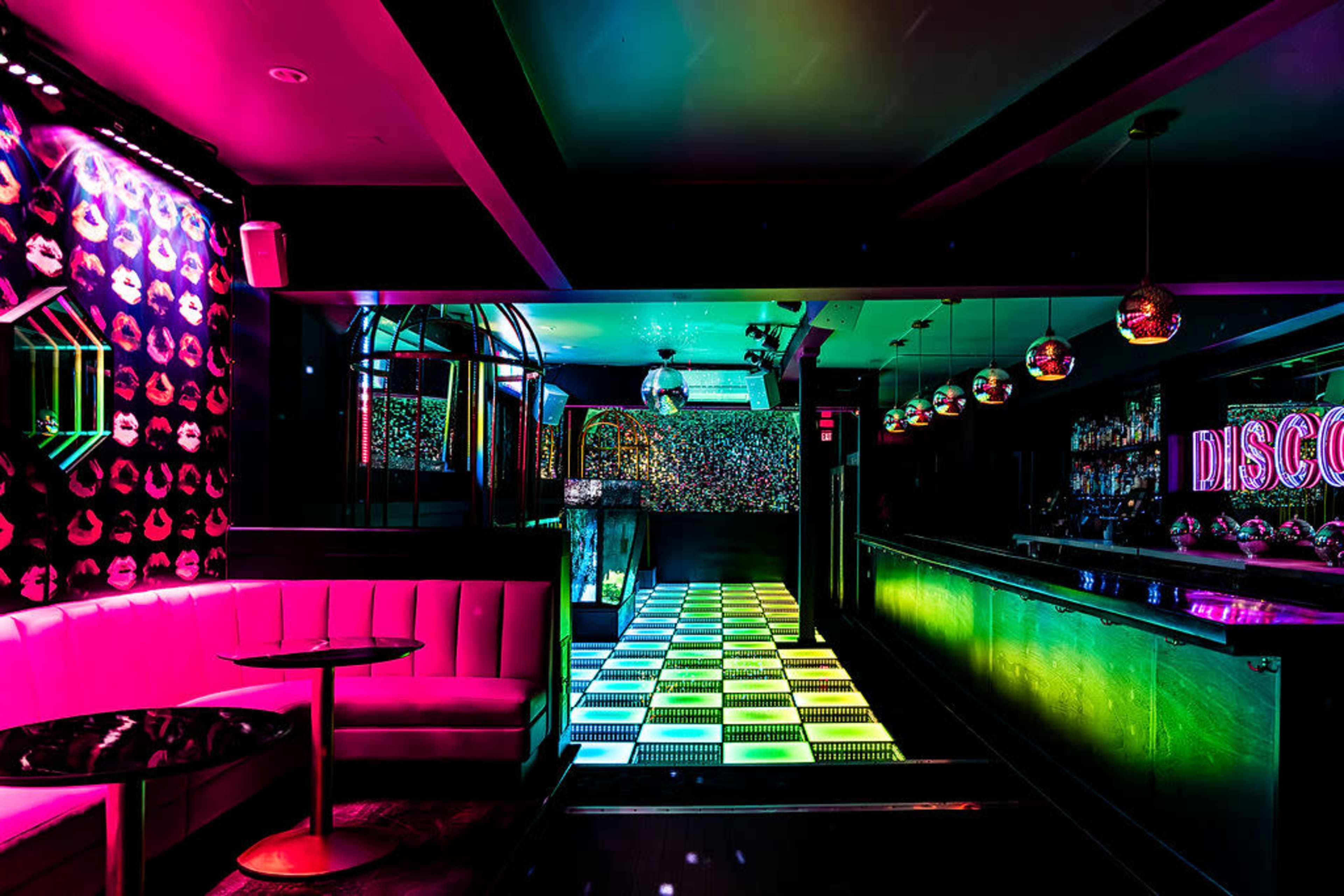 A brightly lit disco bar features a patterned dance floor, colorful neon lighting, and plush seating along one wall.
