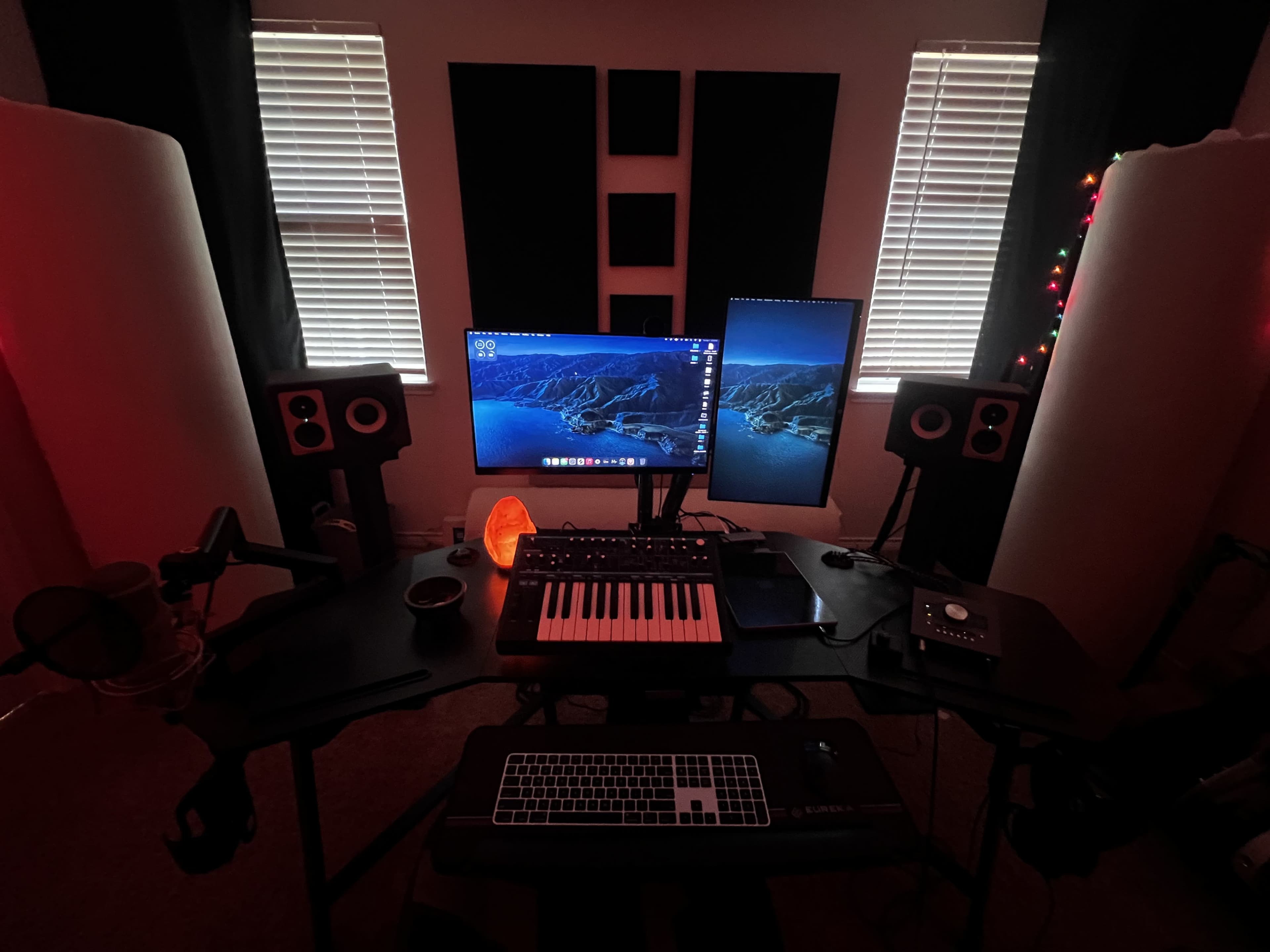 A home music studio features two monitors, a keyboard, speakers, and a microphone, set against a backdrop of black curtains and ambient lighting.