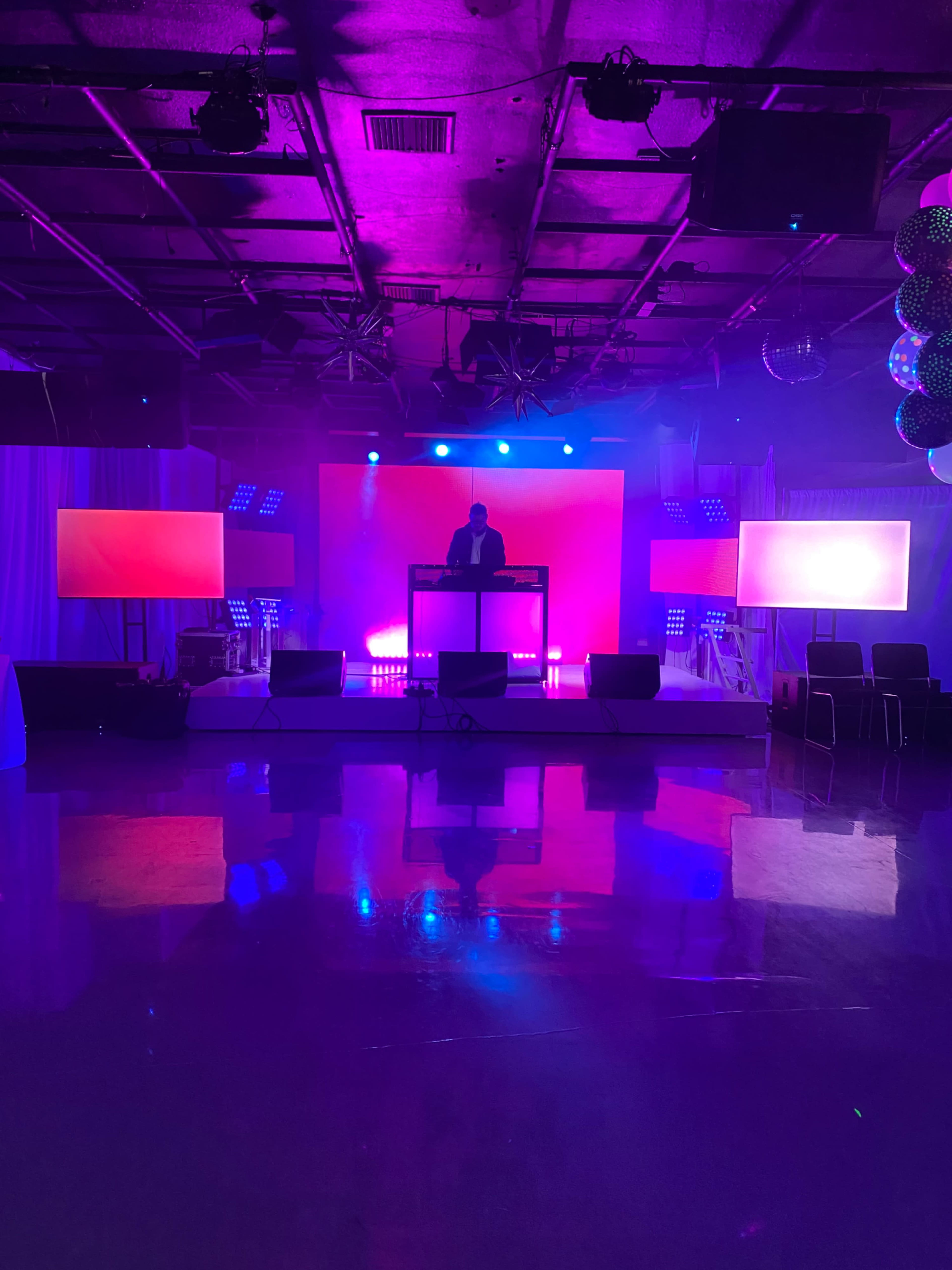A DJ stands at a booth surrounded by colorful lights and screens in a dimly lit event space.
