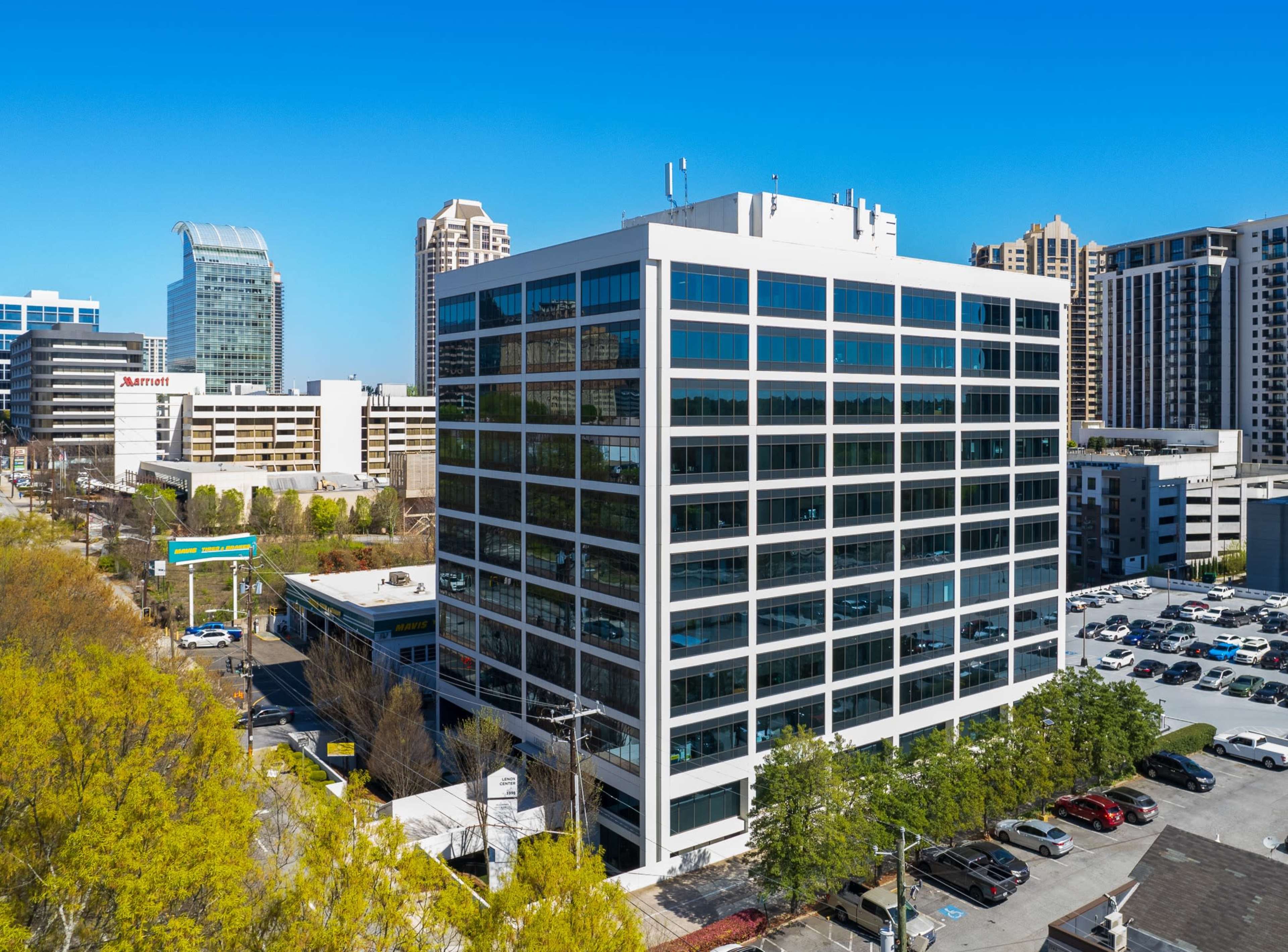 The image shows a modern office building with large glass windows, surrounded by other commercial buildings and a parking lot filled with cars.