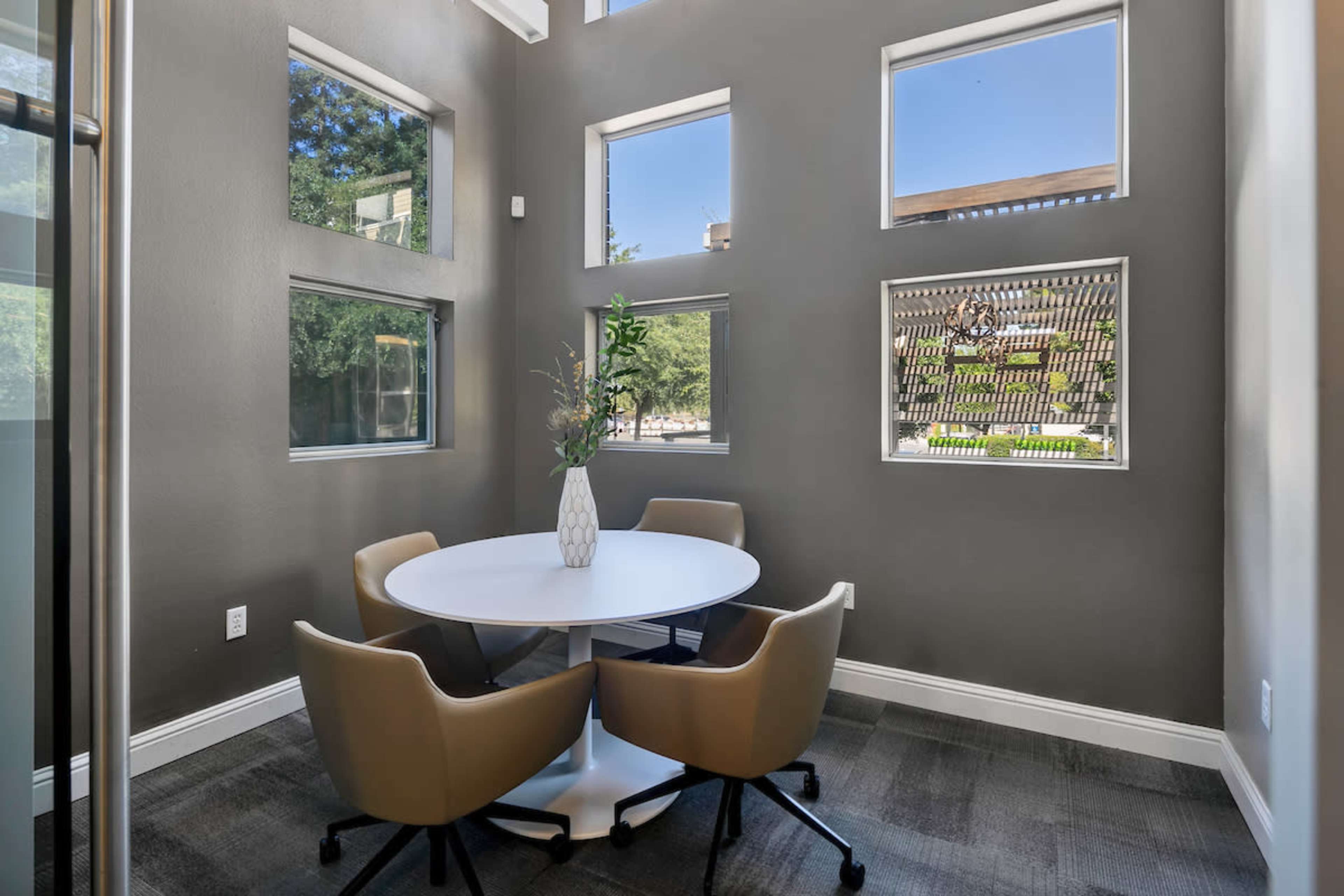 A small meeting room features a round table with four beige chairs, surrounded by large windows letting in natural light.