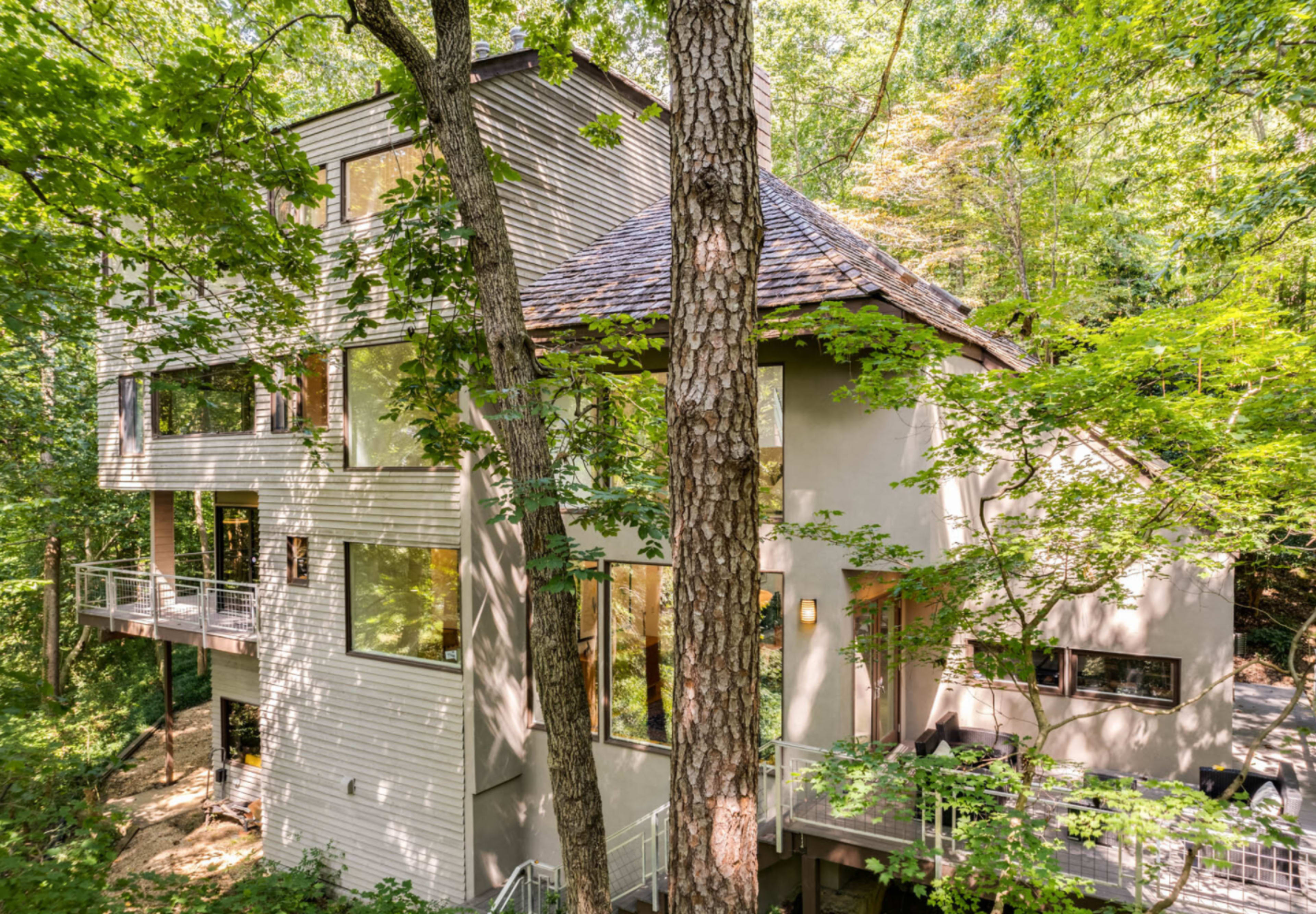 The image shows a multi-story modern house nestled among trees in a wooded area.