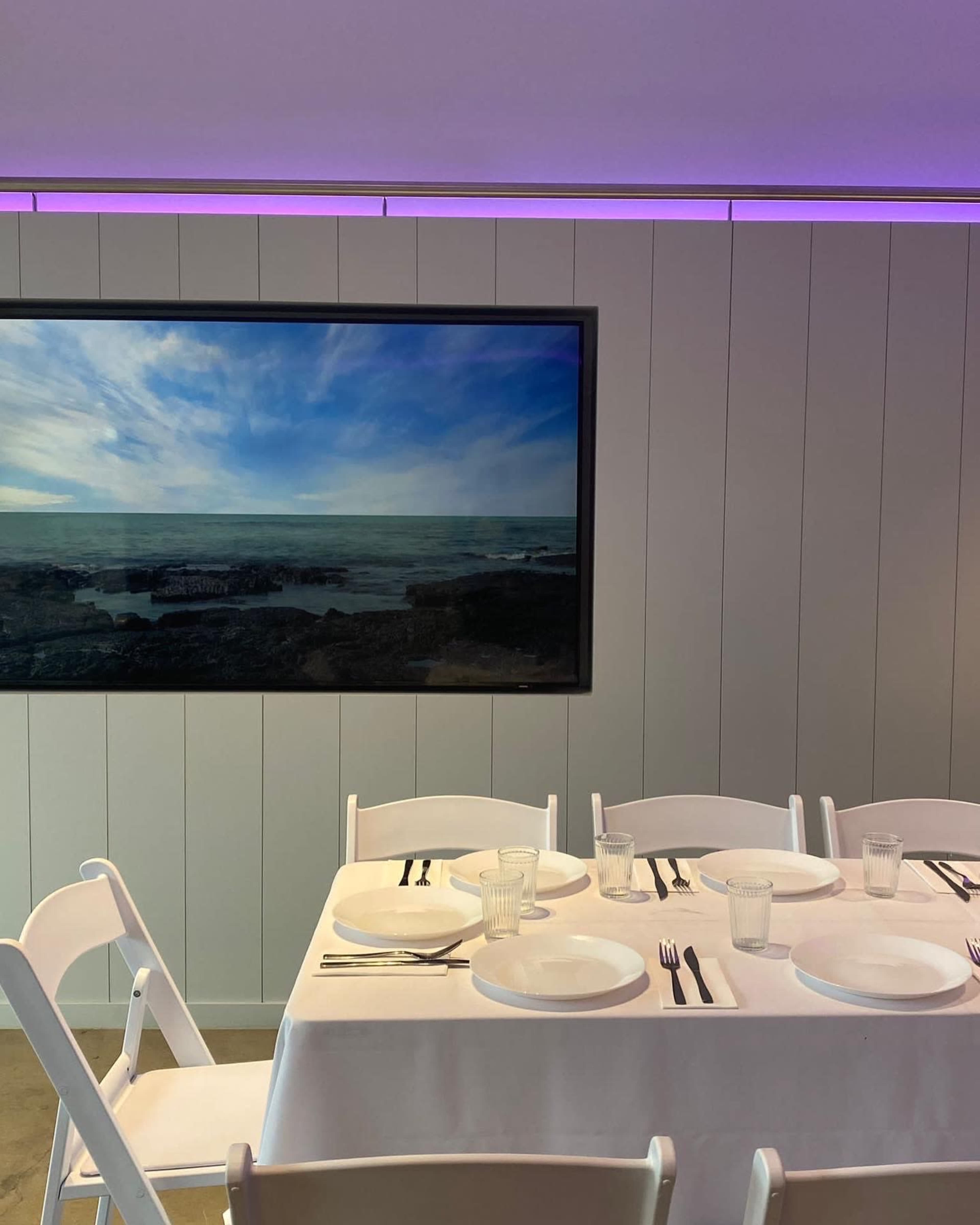 A dining table is set with white plates and utensils in front of a large framed landscape painting, with purple lighting above.