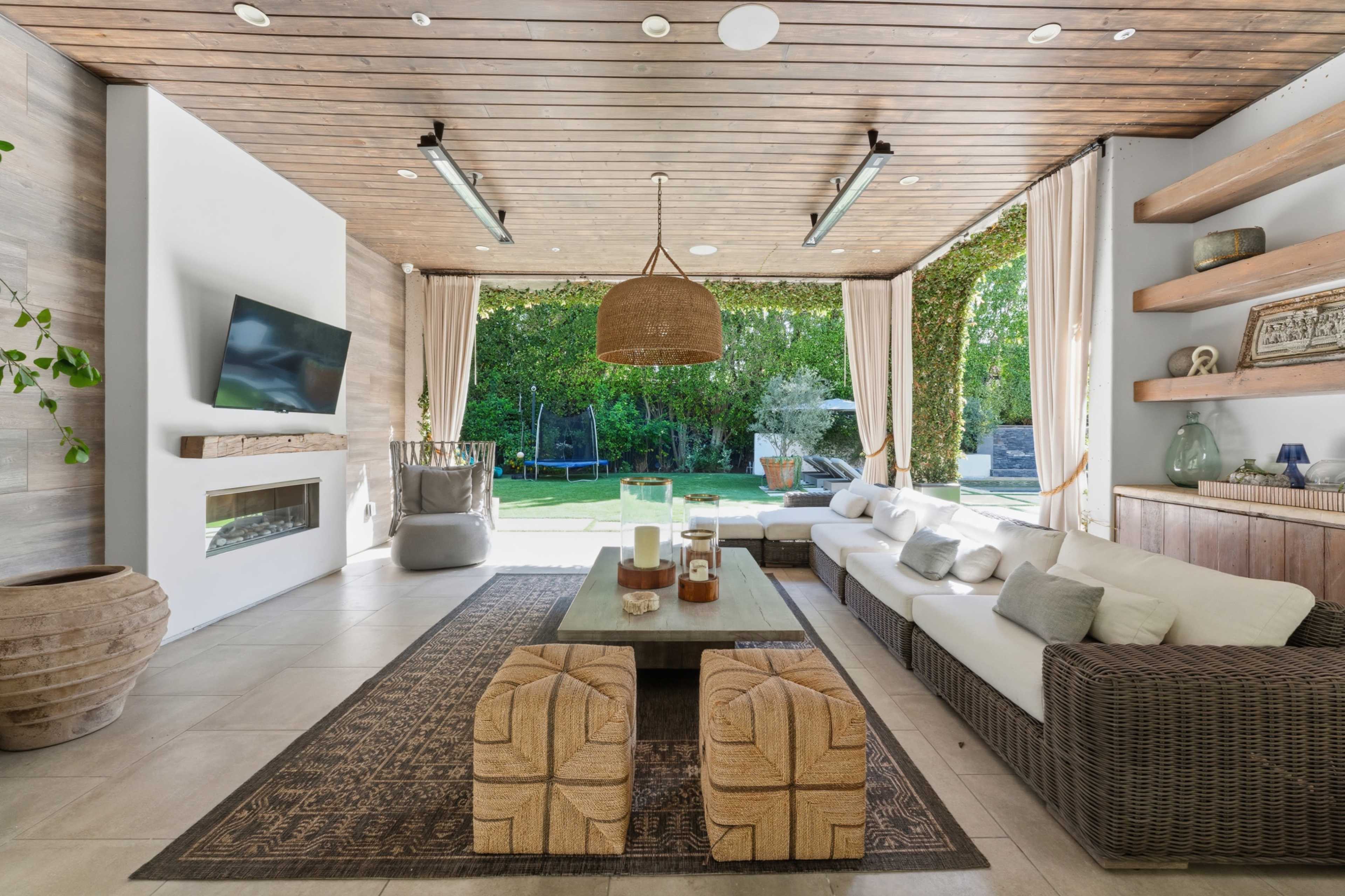 The image shows a modern outdoor living space with a large sectional sofa, a rectangular coffee table, and a television mounted on the wall, featuring sliding glass doors that open to a green backyard.