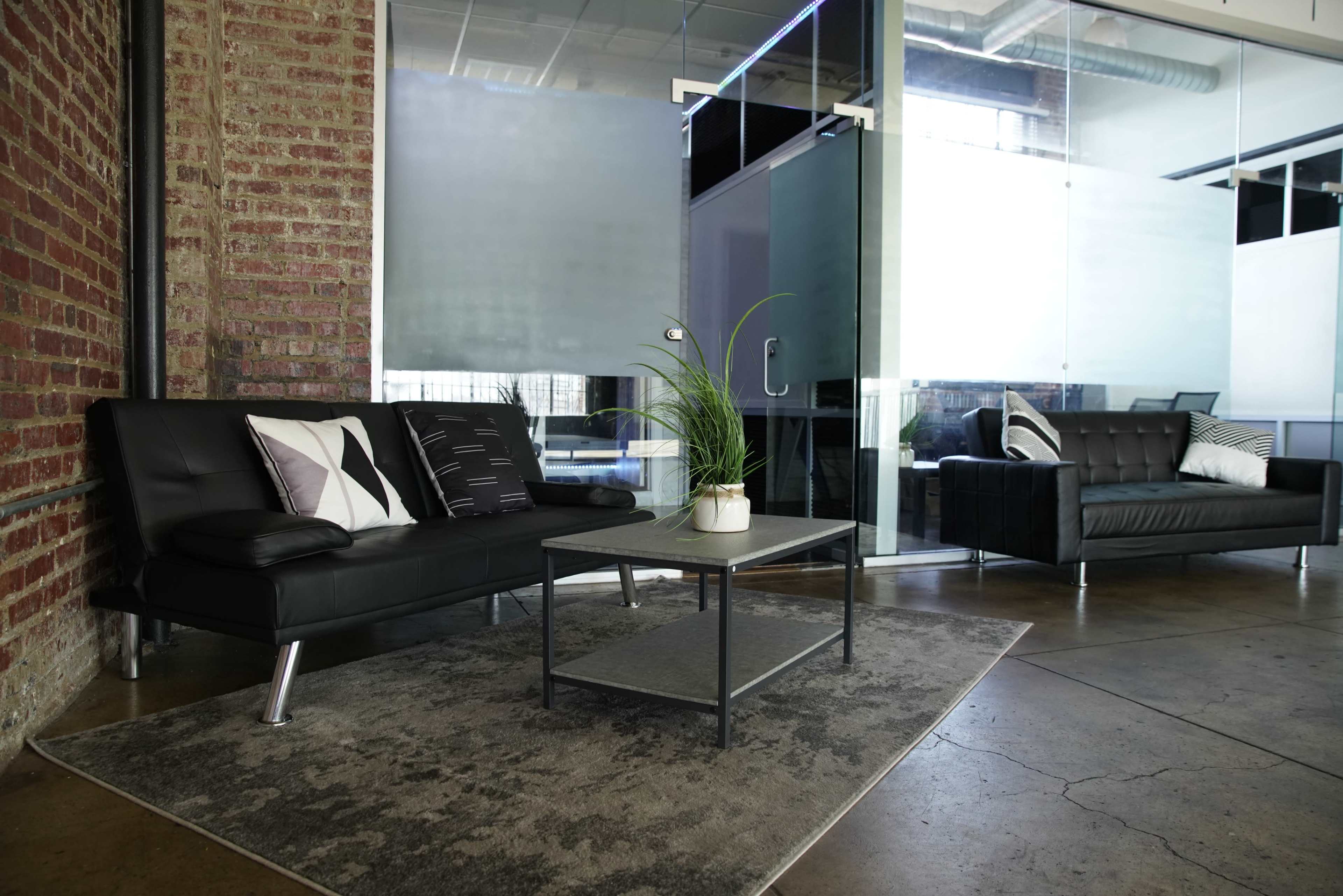 A modern office lounge area featuring black leather sofas, a glass partition, and a coffee table with a potted plant on a patterned rug.