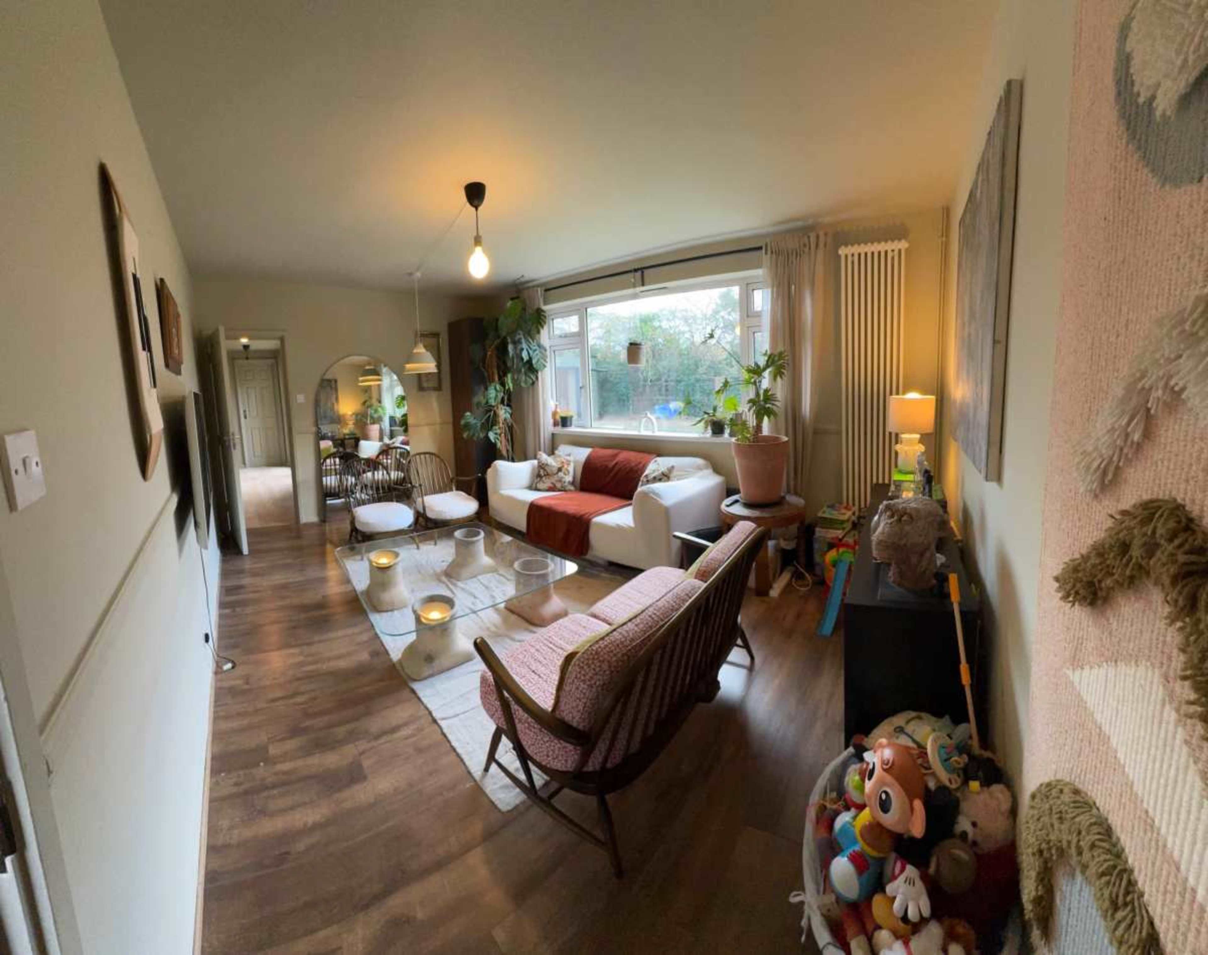 The image shows a living room with a white sofa, a wooden coffee table, and a variety of plants, toys, and decor items.