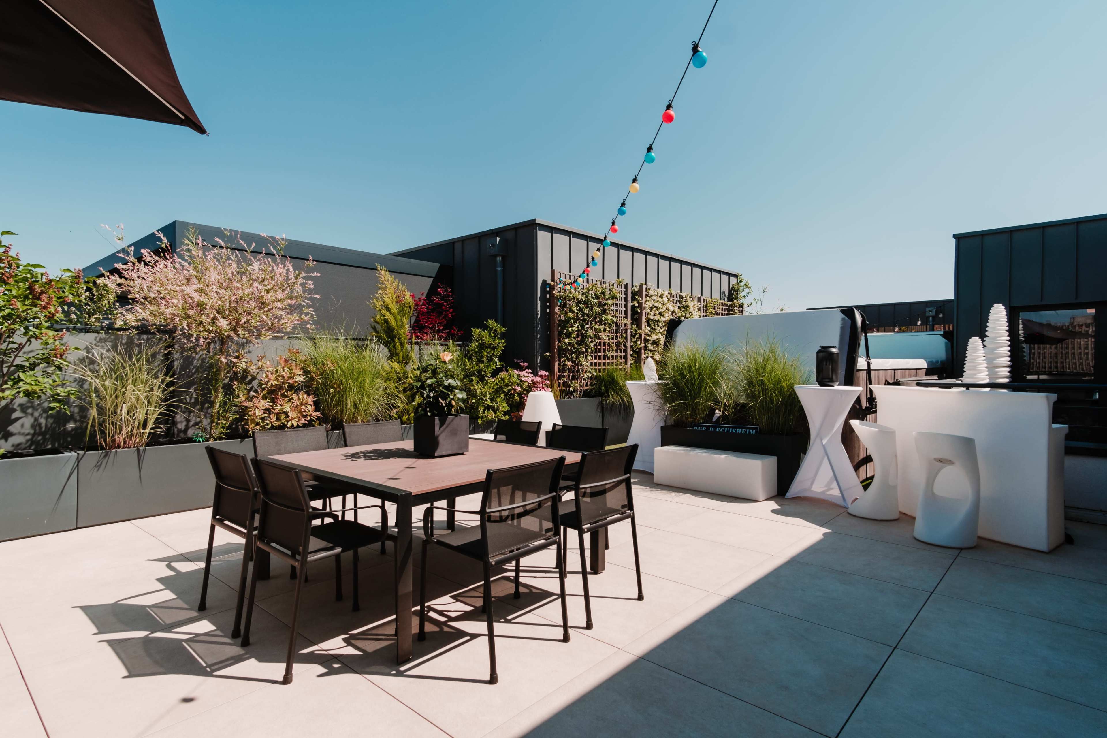 A modern rooftop terrace features a wooden dining table surrounded by black chairs, decorated with lush greenery and colorful hanging lights under a clear blue sky.