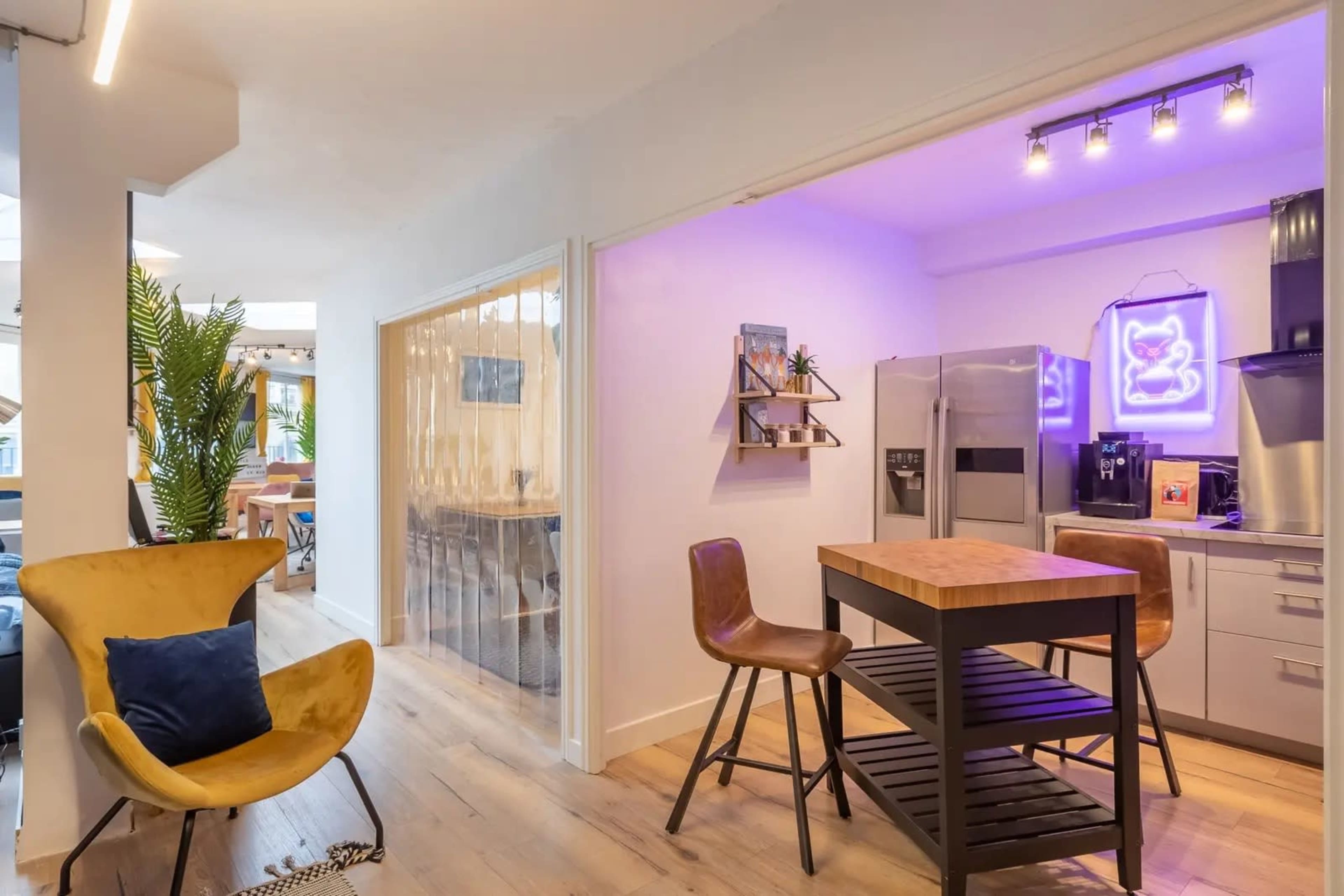 The image shows a modern kitchen and dining area with a wooden table, metal chairs, and a colorful neon light.