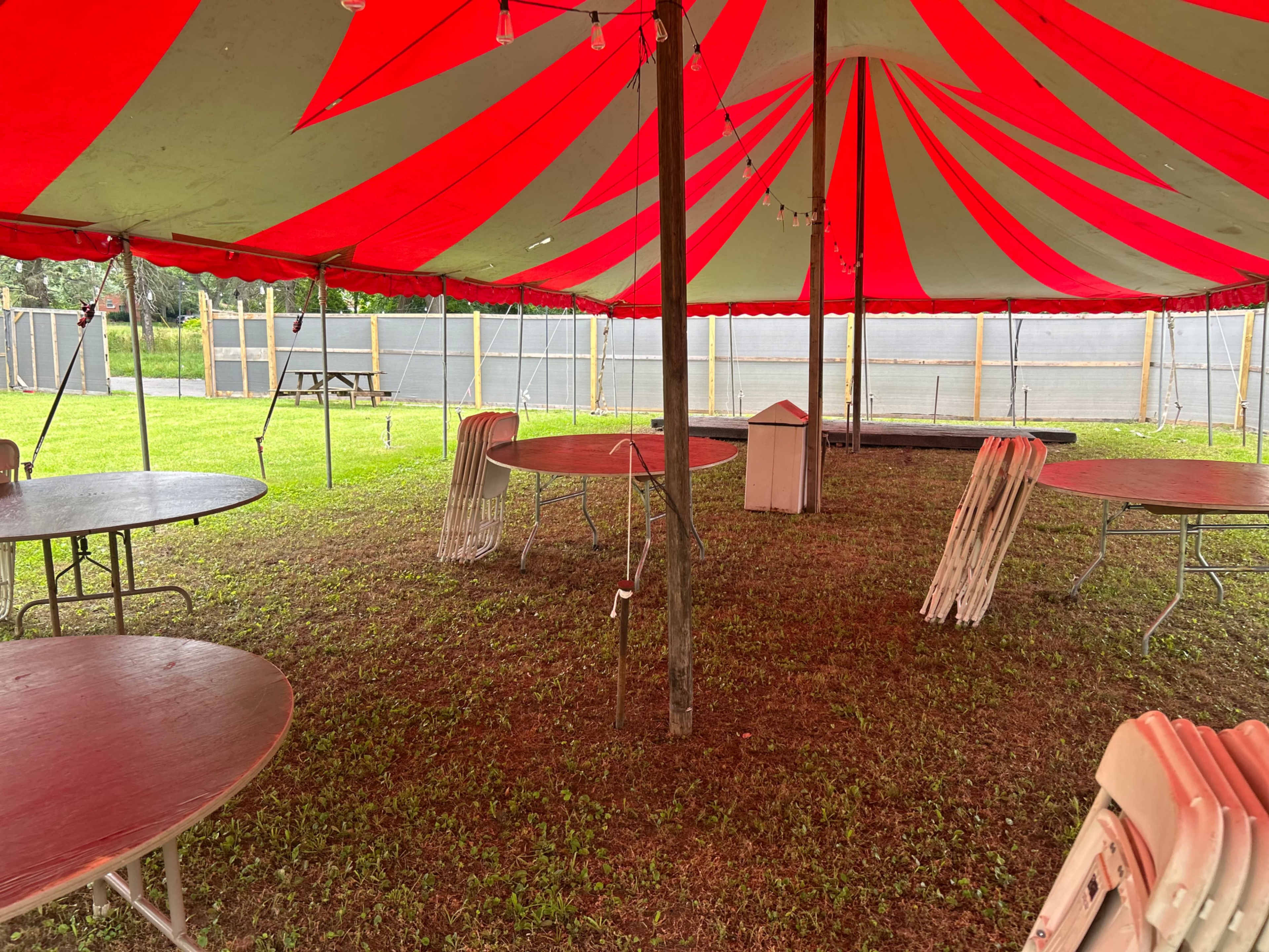 A circus tent with red and white stripes is set up on grassy ground, featuring empty tables and chairs arranged beneath it.