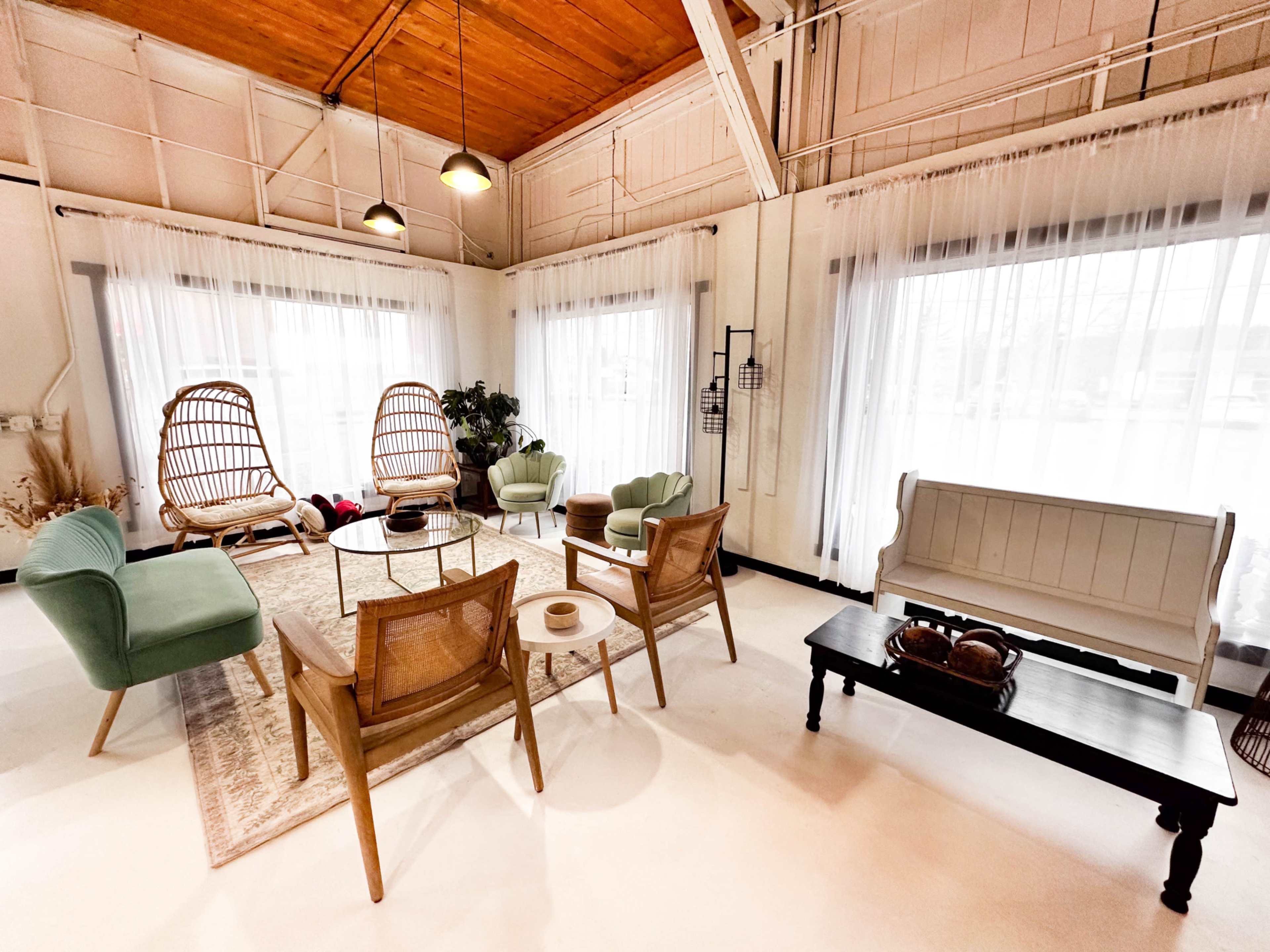 The image shows a modern, airy living space with a mix of seating options, including two wicker chairs, a green armchair, and a wooden bench, surrounded by large windows adorned with sheer white curtains.