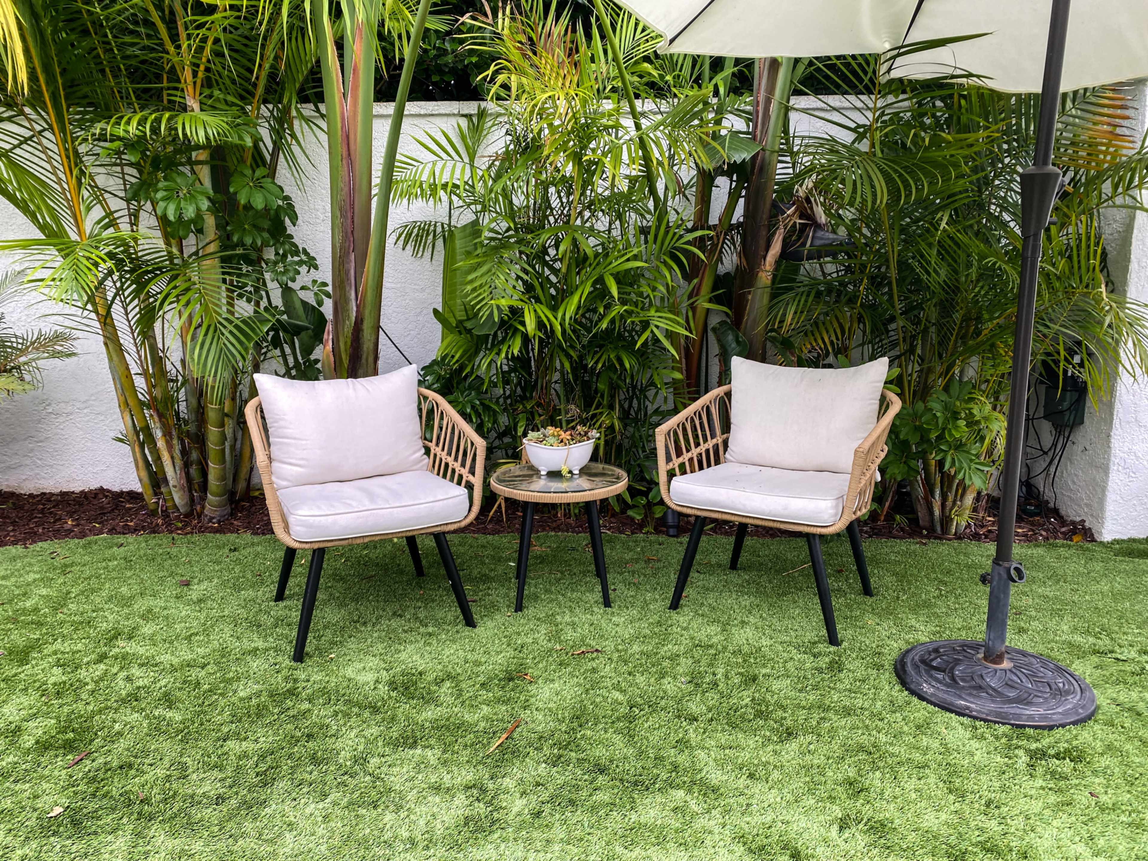 Two wicker chairs with light cushions sit beside a small table on artificial grass, surrounded by lush greenery.