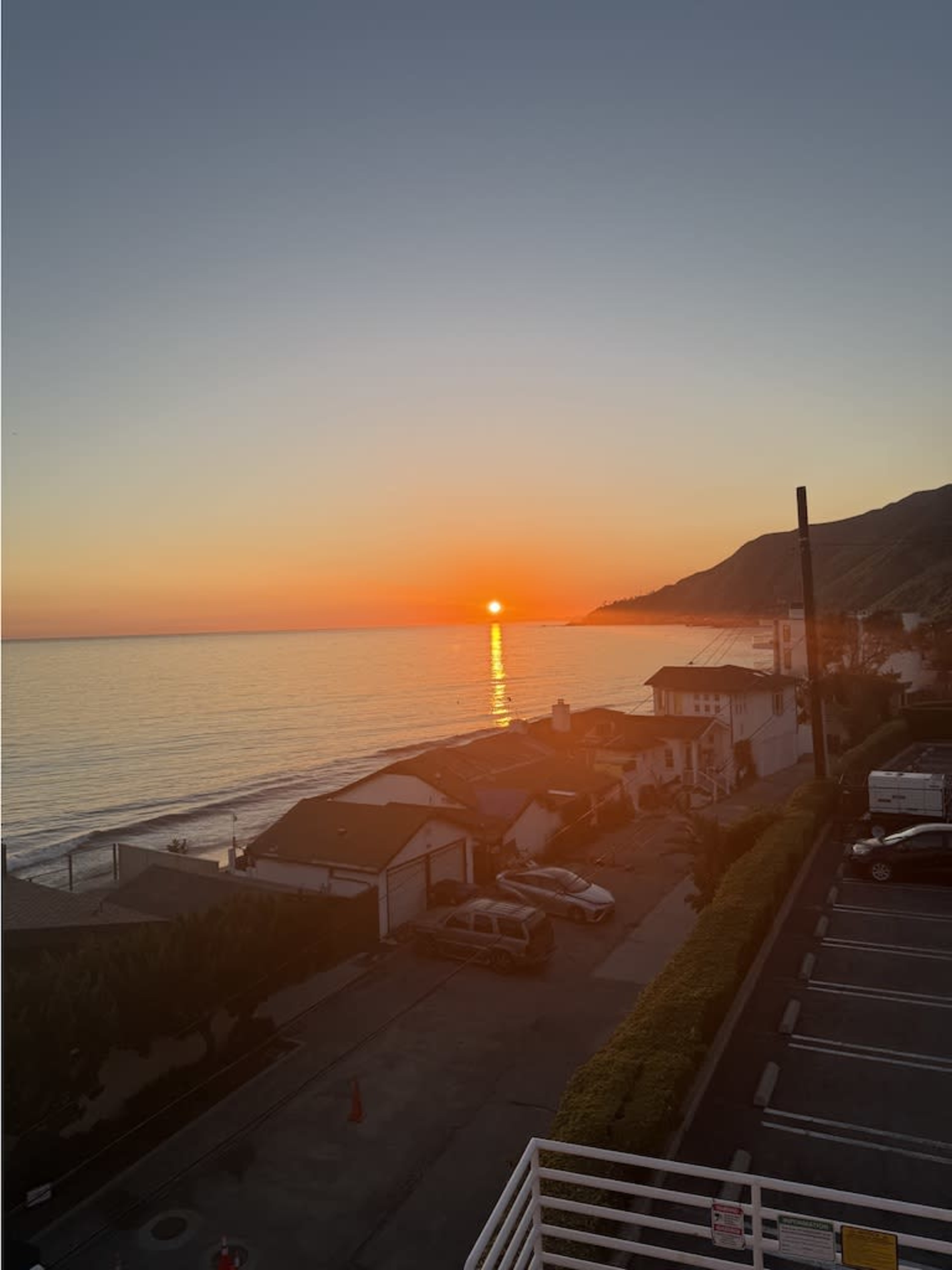 The image shows a sunset over the ocean, with the sun partially dipped below the horizon, casting a warm glow across the water and illuminating the shoreline lined with houses.