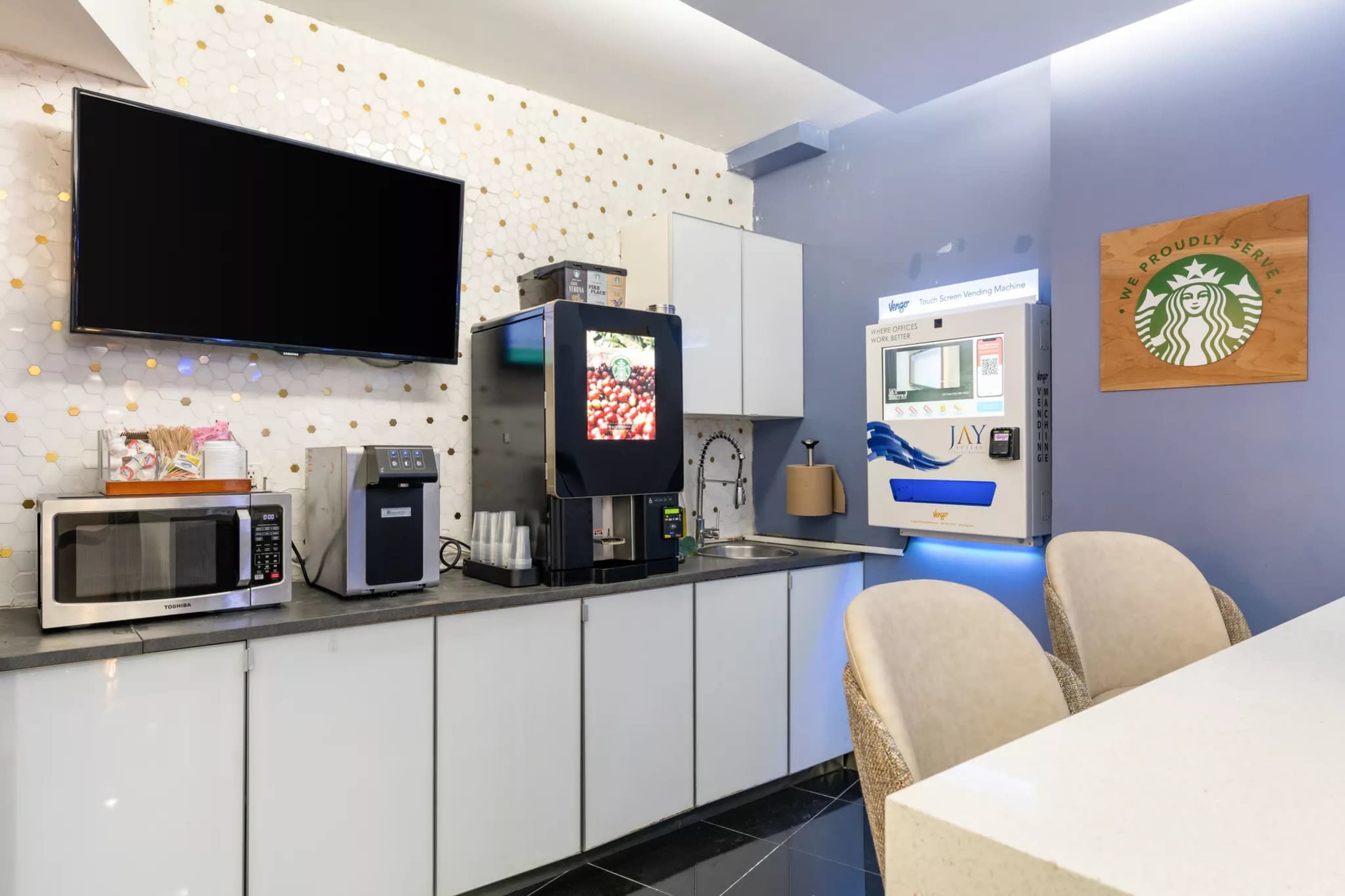The image shows a modern kitchenette area featuring a microwave, a beverage dispenser, a coffee machine, and a wall-mounted TV.