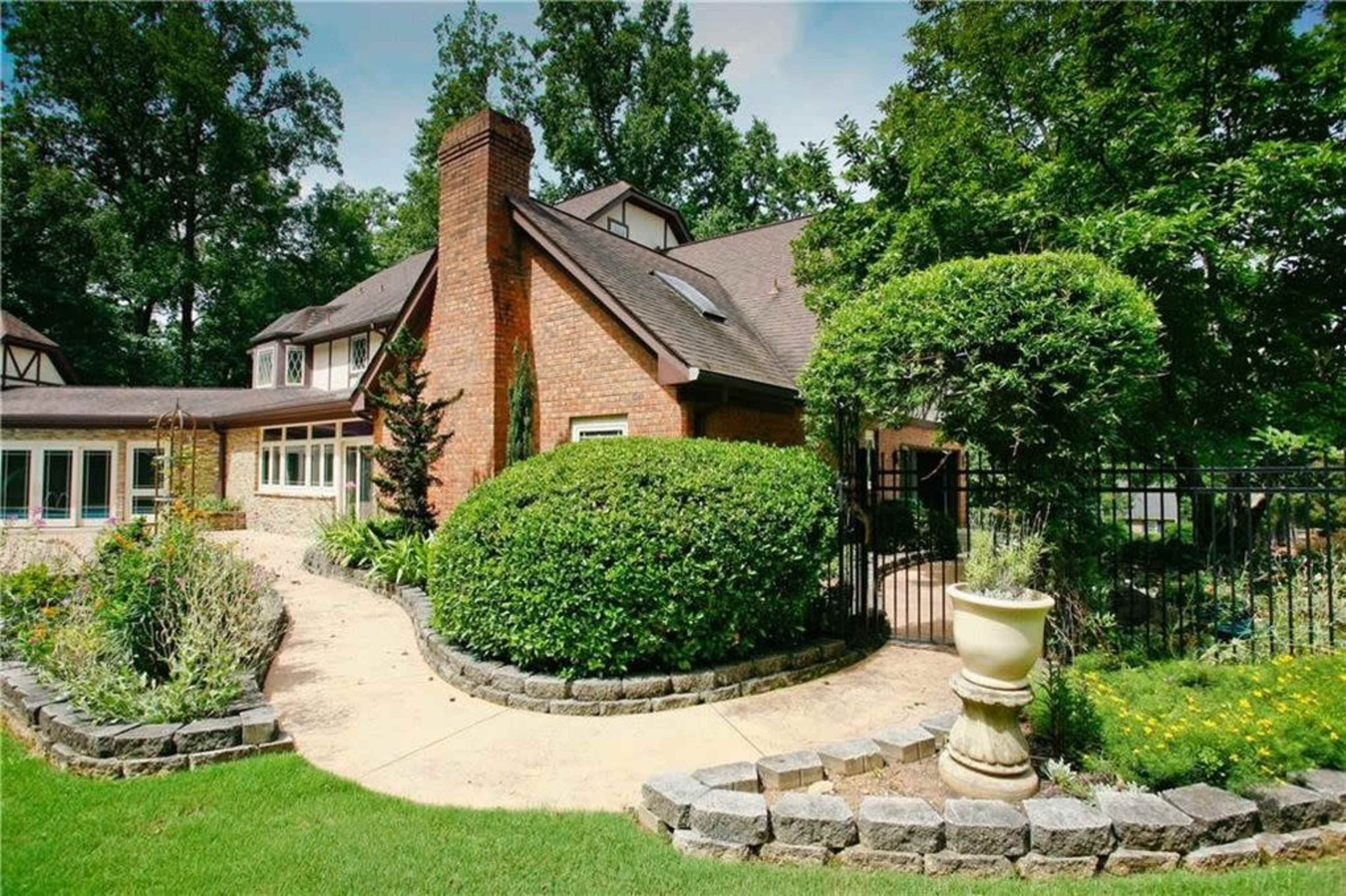 A brick home with a sloped roof is surrounded by landscaped gardens and a stone pathway leading to a gated area.