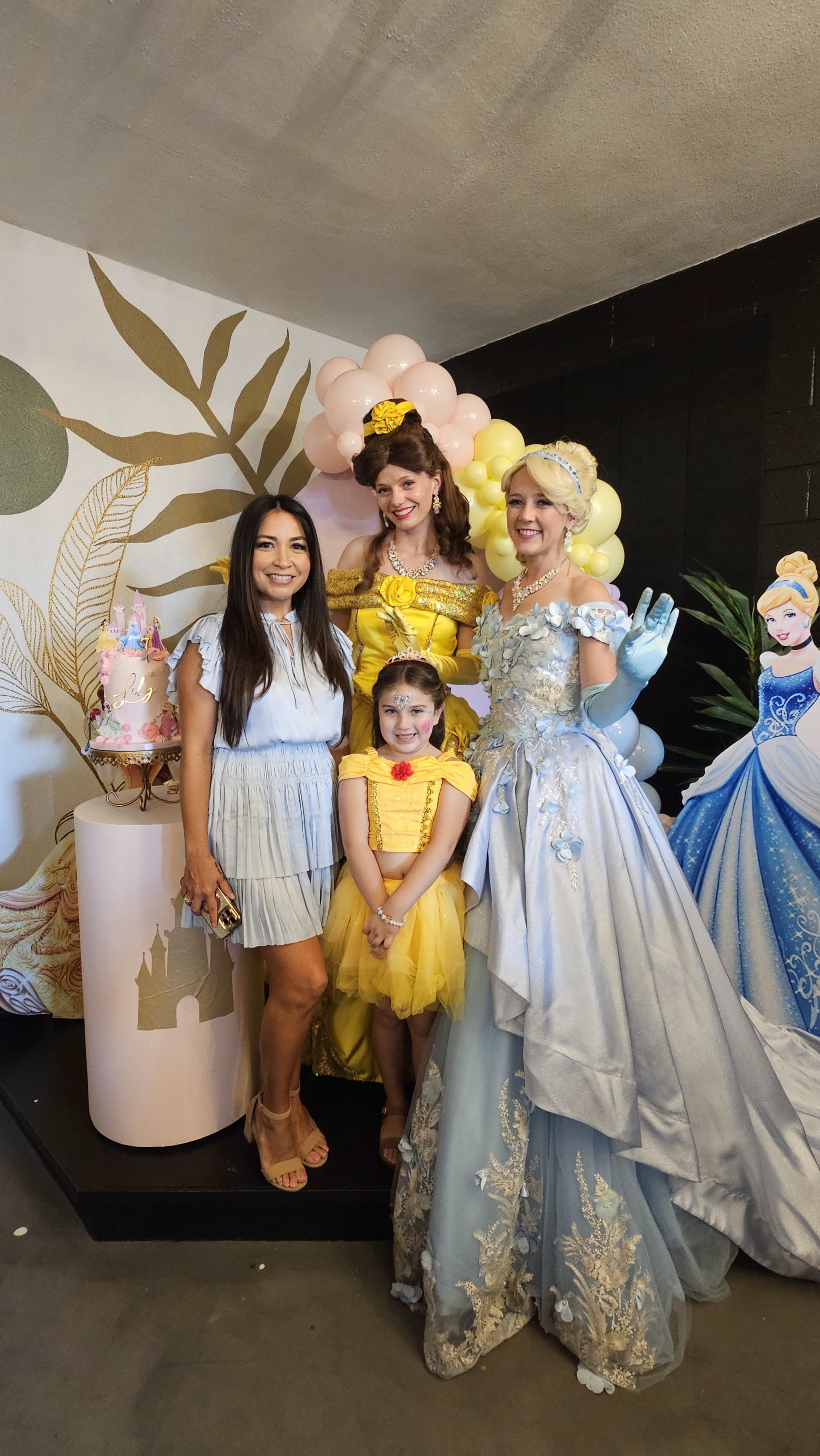 A woman and a girl in yellow dresses pose with two women dressed as princesses at a themed party.