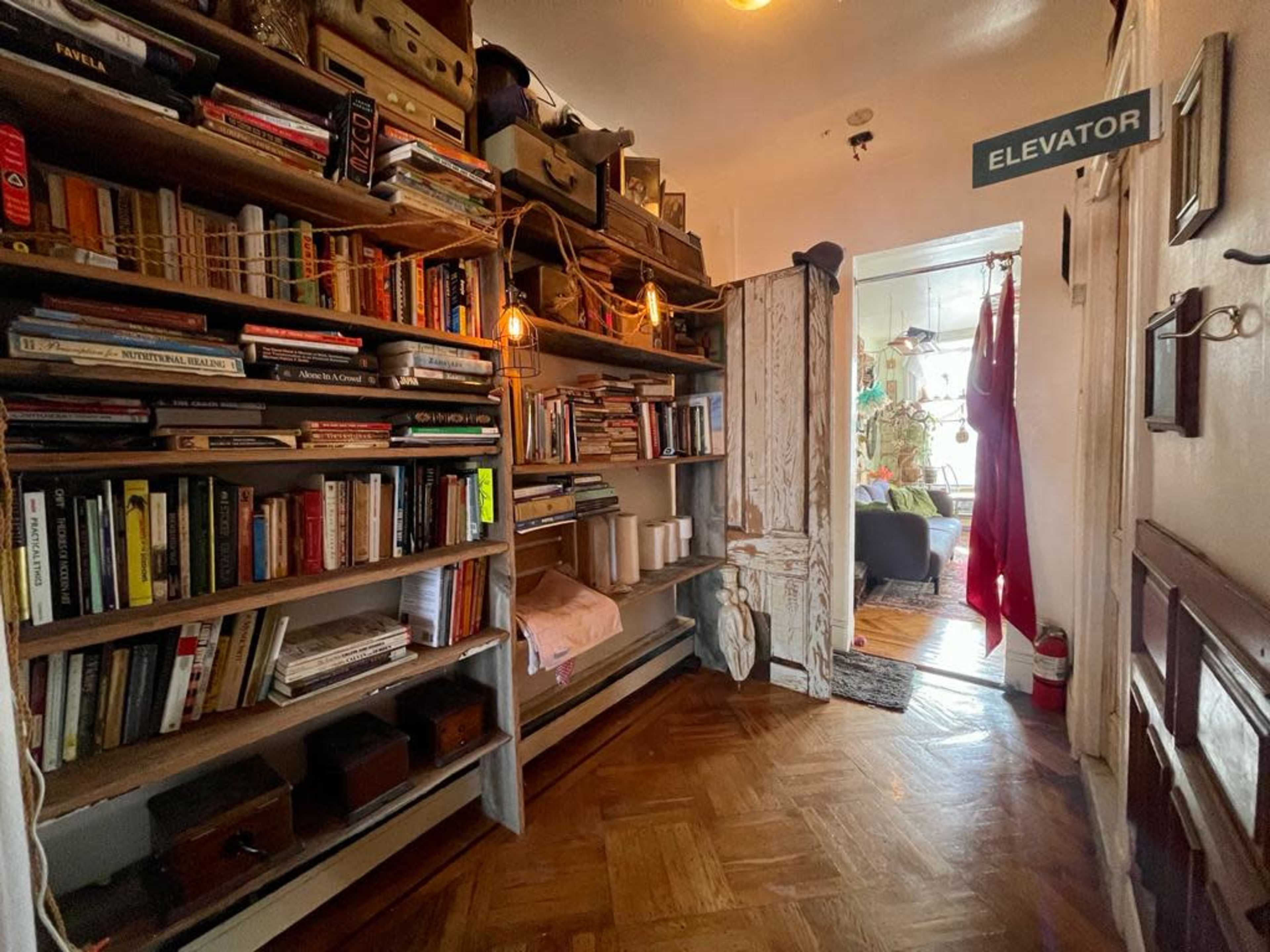 A narrow hallway features shelves filled with books, a vintage clothes rack, and a sign indicating the location of an elevator.