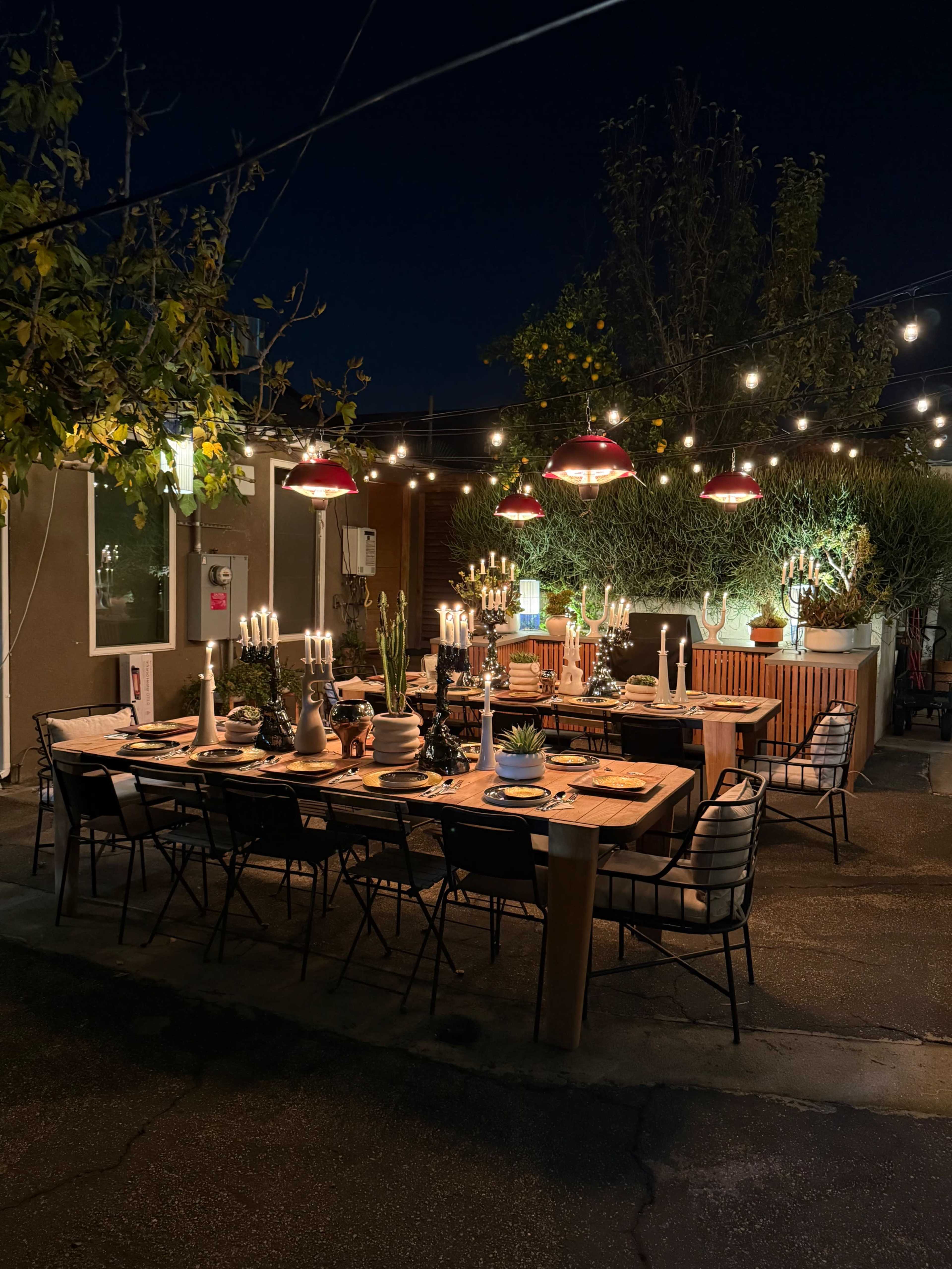 A dining table is set outdoors at night, adorned with candles and surrounded by string lights, with greenery in the background.