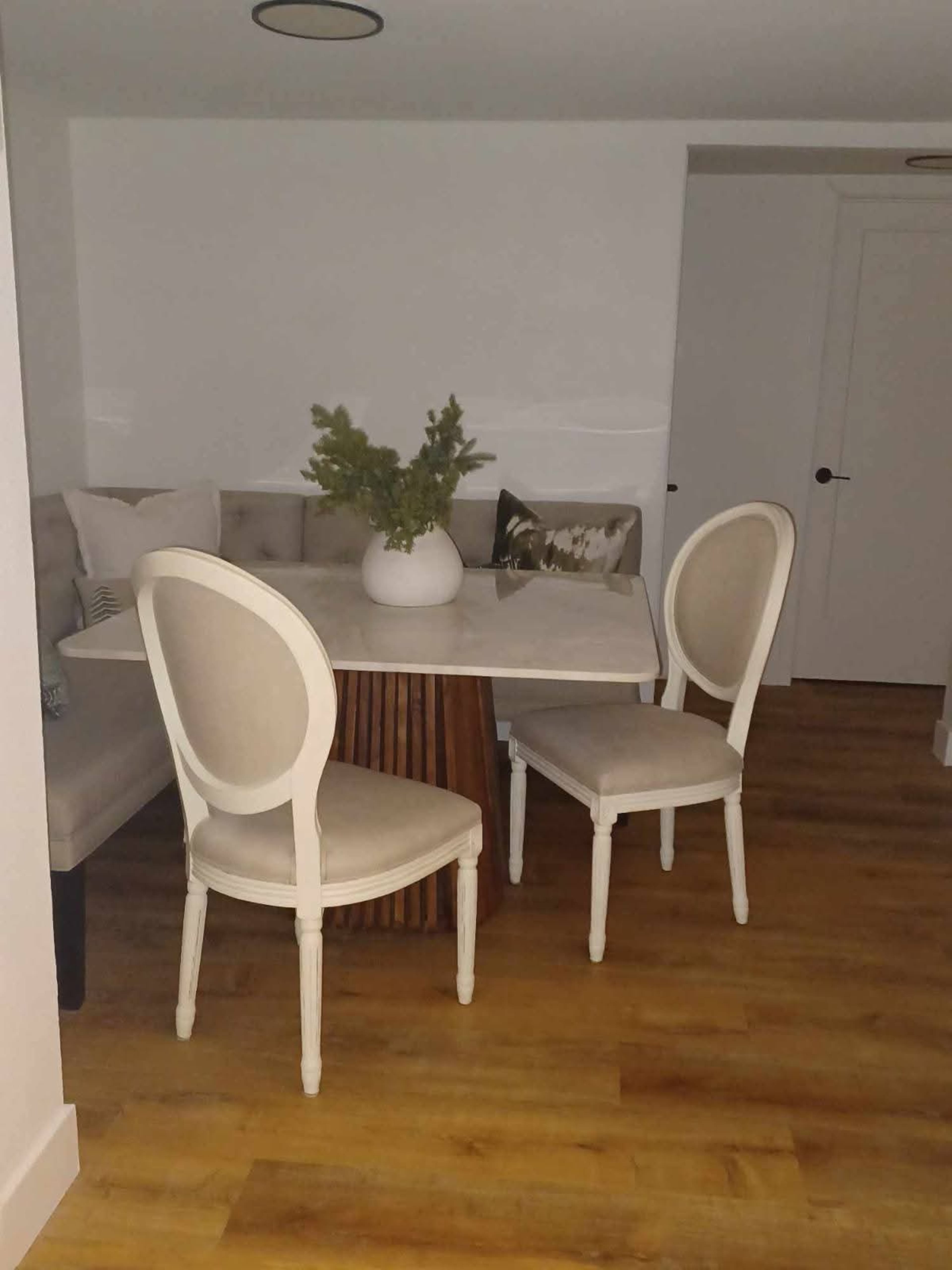 A small dining area features a round table with a decorative vase and two chairs near a cushioned bench against the wall.
