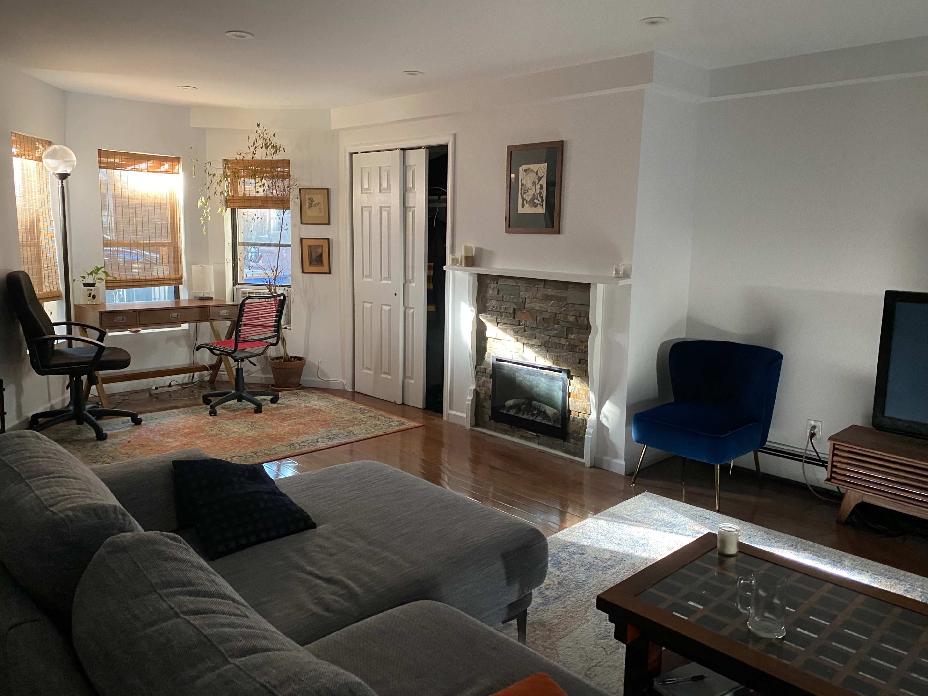 A living room with a gray sectional sofa, a wooden coffee table, a stone fireplace, a work desk with a chair, and a blue armchair, illuminated by natural light.