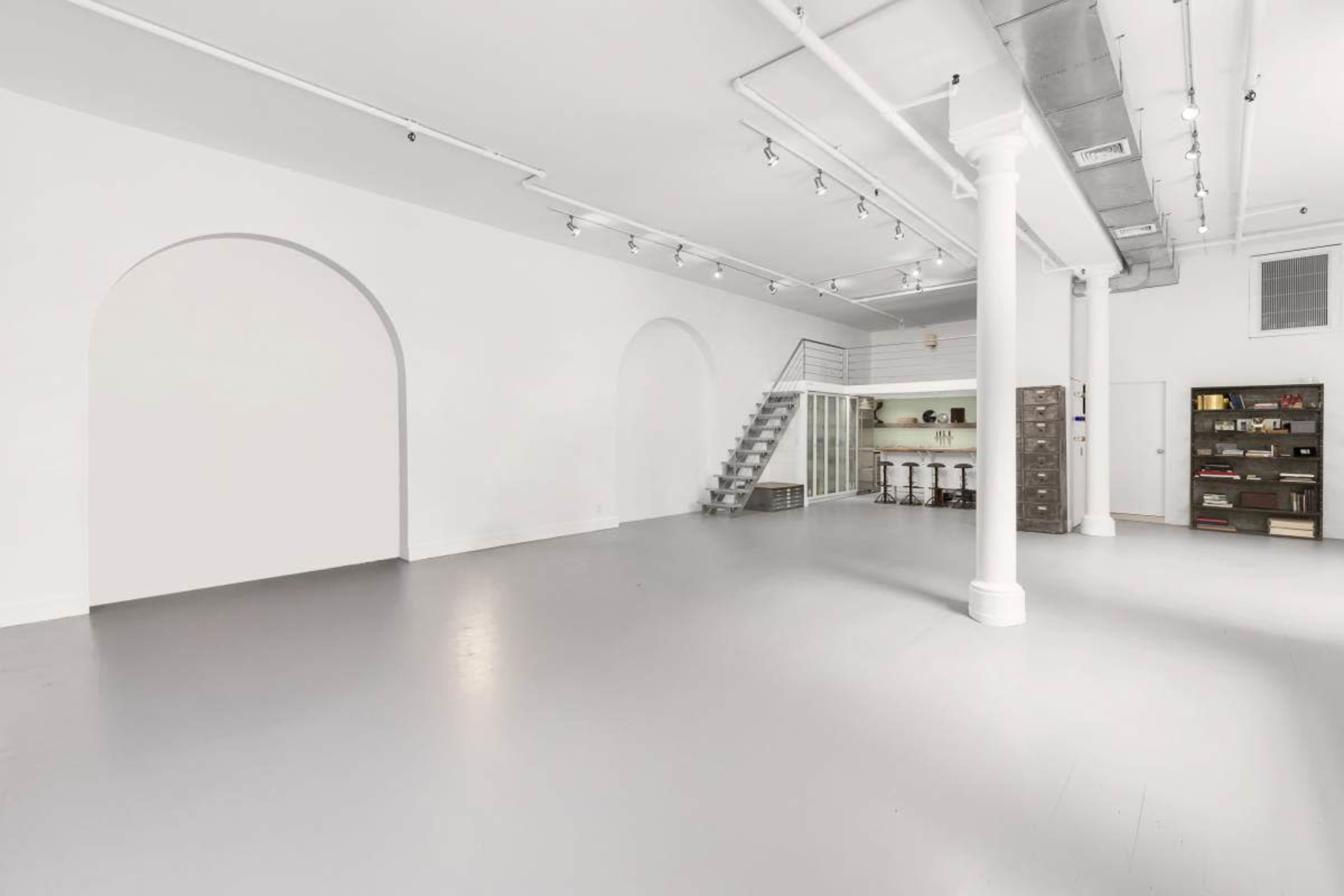 The image shows an expansive, minimally decorated interior space with light gray flooring and white walls, featuring a staircase leading to an upper level.